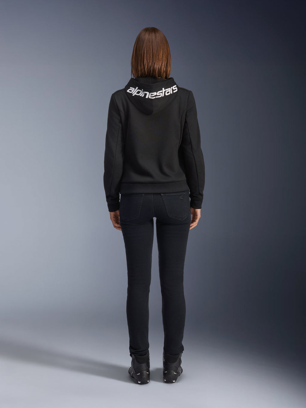 Daisy V3 Women's Riding Denim, pantalones de moto para mujer de corte slim, color negro, vista trasera que muestra la cintura alta y la silueta femenina, combinados con una sudadera con capucha negra de Alpinestars y botas de moto negras, equipamiento de conducción urbana casual