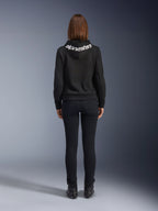 Daisy V3 Women's Riding Denim, pantalones de moto para mujer de corte slim, color negro, vista trasera que muestra la cintura alta y la silueta femenina, combinados con una sudadera con capucha negra de Alpinestars y botas de moto negras, equipamiento de conducción urbana casual