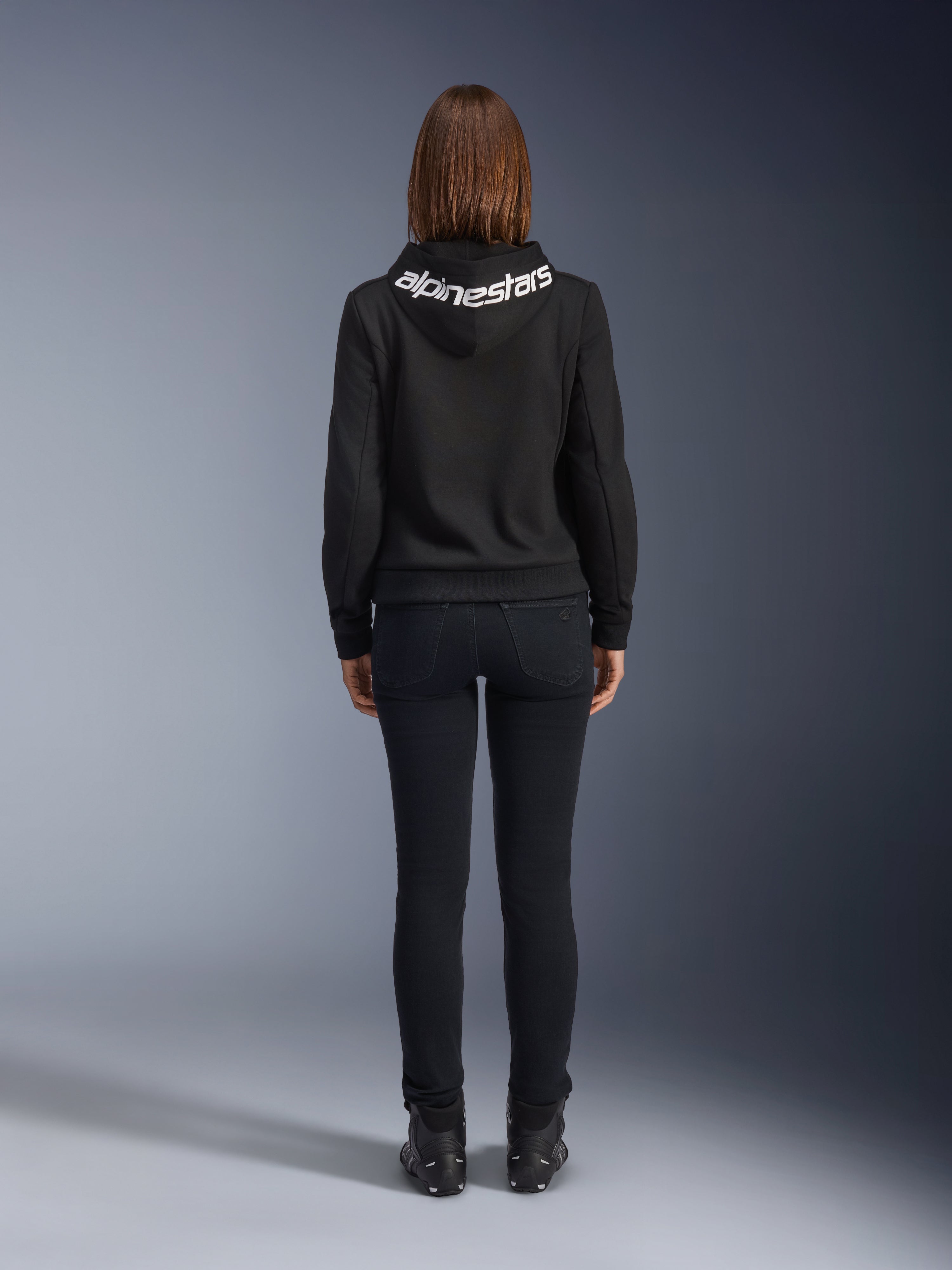 Daisy V3 Women's Riding Denim, pantalones de moto para mujer de corte slim, color negro, vista trasera que muestra la cintura alta y la silueta femenina, combinados con una sudadera con capucha negra de Alpinestars y botas de moto negras, equipamiento de conducción urbana casual