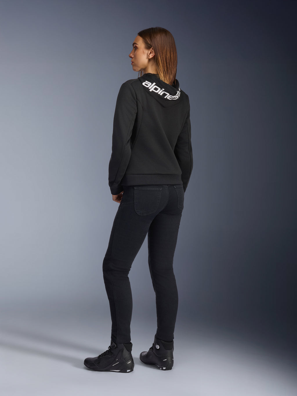 Alpinestars Daisy V3 Women's Riding Denim, pantalones de moto para mujer, negro, vista trasera que muestra el ajuste entallado y el talle alto, combinado con una sudadera Alpinestars negra y botas de moto para un look motero urbano