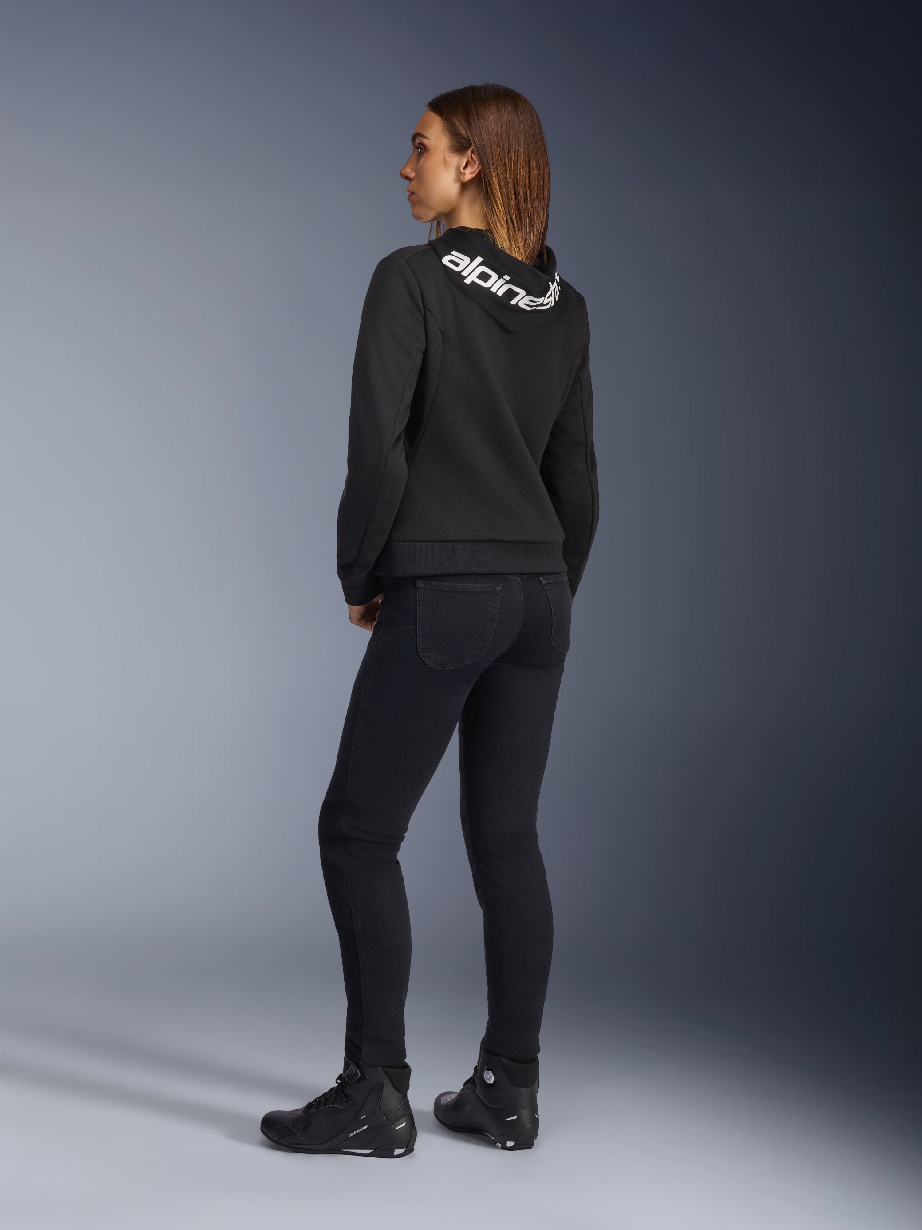 Alpinestars Daisy V3 Women's Riding Denim, pantalones de moto para mujer, negro, vista trasera que muestra el ajuste entallado y el talle alto, combinado con una sudadera Alpinestars negra y botas de moto para un look motero urbano