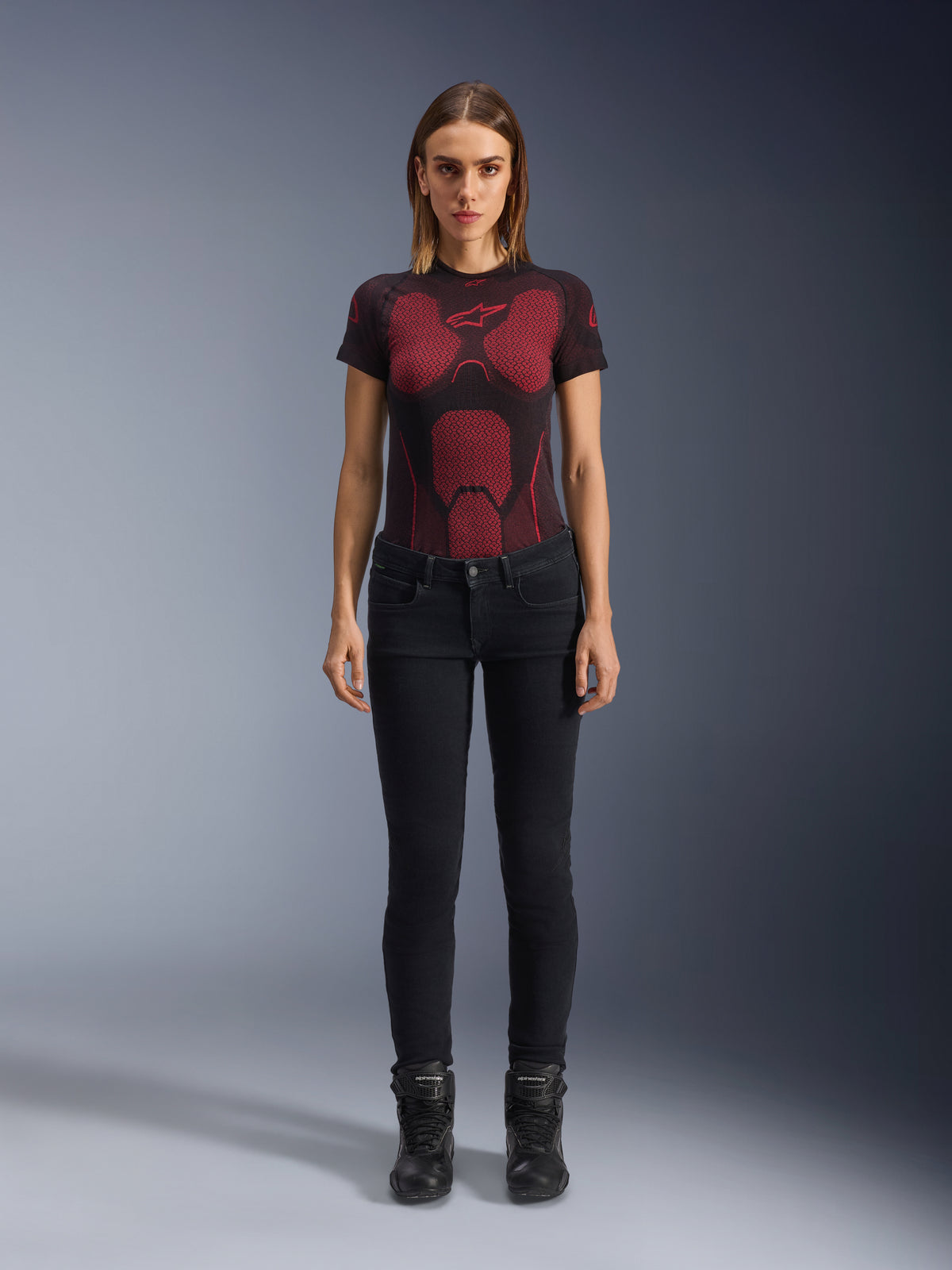 Alpinestars Daisy V3 Women's Riding Denim, pantalones de moto para mujer de corte ajustado en color Rinse Black, lucidos por una modelo con una camiseta técnica de patrón rojo y negro y botas de moto negras, equipamiento de conducción urbana casual con silueta femenina y diseño de talle alto.