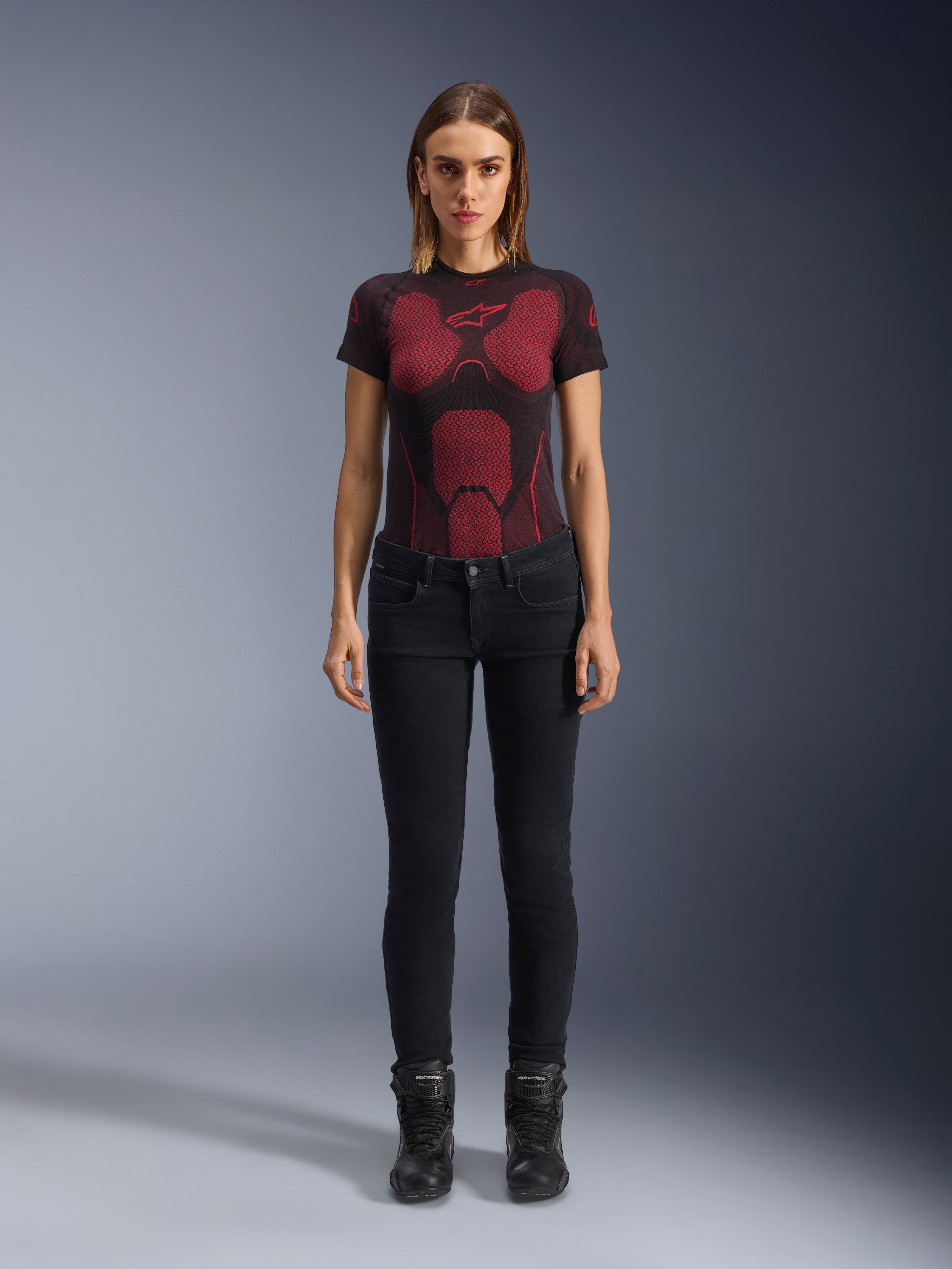Alpinestars Daisy V3 Women's Riding Denim, pantalones de moto para mujer de corte ajustado en color Rinse Black, lucidos por una modelo con una camiseta técnica de patrón rojo y negro y botas de moto negras, equipamiento de conducción urbana casual con silueta femenina y diseño de talle alto.