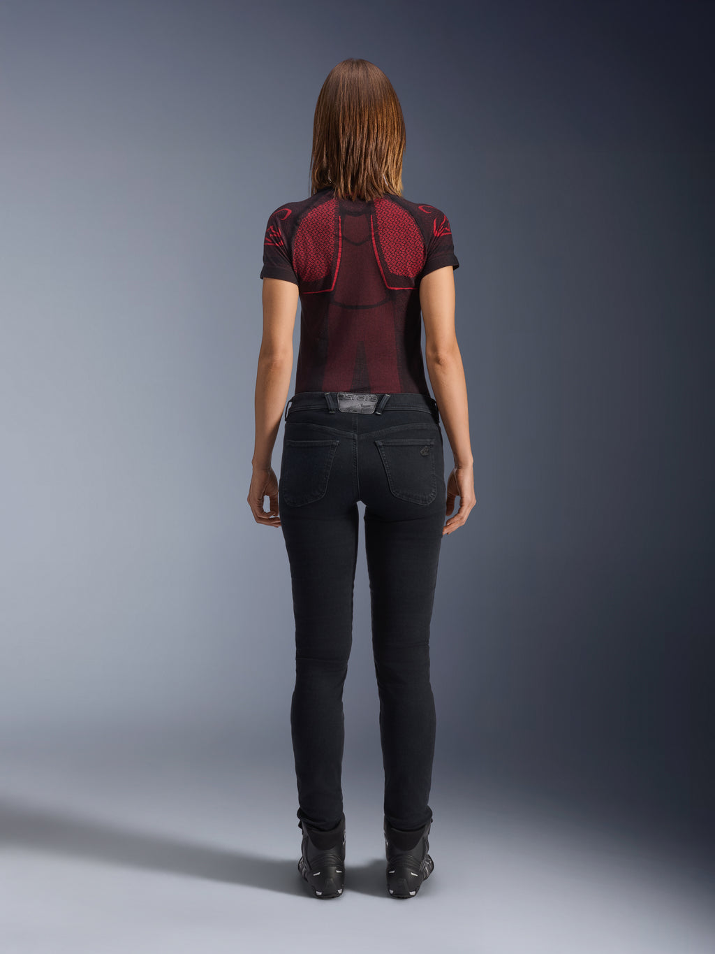Alpinestars Daisy V3 Women's Riding Denim, Rinse Black, pantalones de moto de corte ajustado vistos desde atrás, con diseño de cintura alta y bolsillos traseros, combinados con una camiseta técnica con estampado en rojo y negro y botas negras, equipamiento de moto urbano para mujer