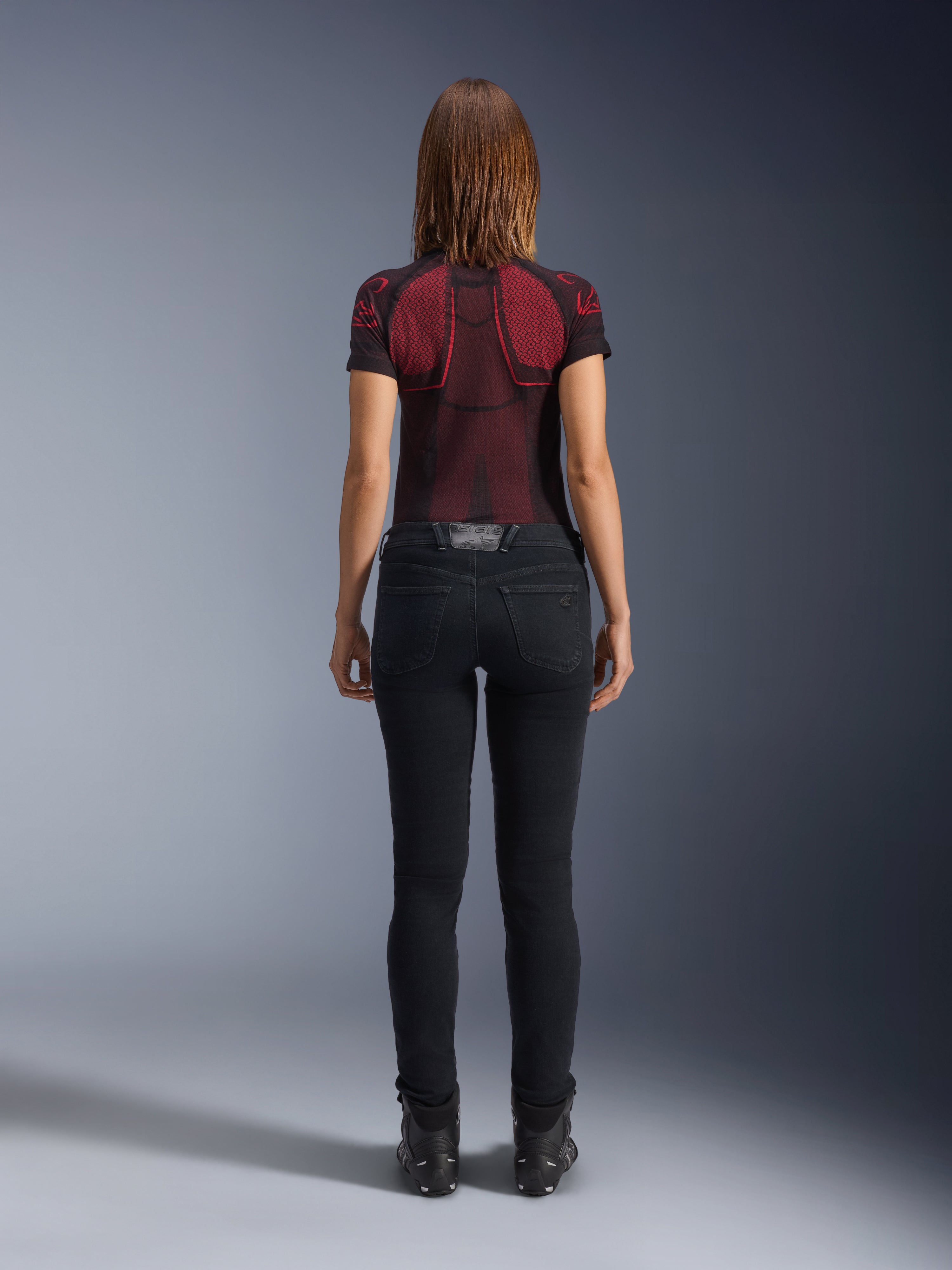 Alpinestars Daisy V3 Women's Riding Denim, Rinse Black, pantalones de moto de corte ajustado vistos desde atrás, con diseño de cintura alta y bolsillos traseros, combinados con una camiseta técnica con estampado en rojo y negro y botas negras, equipamiento de moto urbano para mujer
