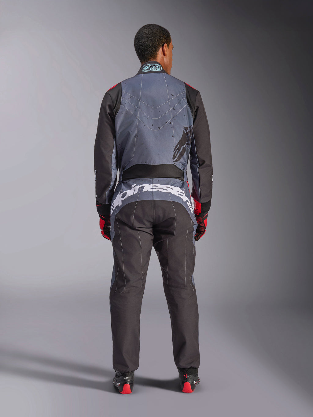 Alpinestars KMX-9 V3 Graphic 6 Suit, mono de karting, negro antracita y rojo, vista trasera que muestra un gran logotipo blanco de Alpinestars en la parte baja de la espalda, parte superior del cuerpo gris con gráficos de salpicaduras negras, brazos y piernas negros, usado con guantes y botas de carreras en rojo y negro.