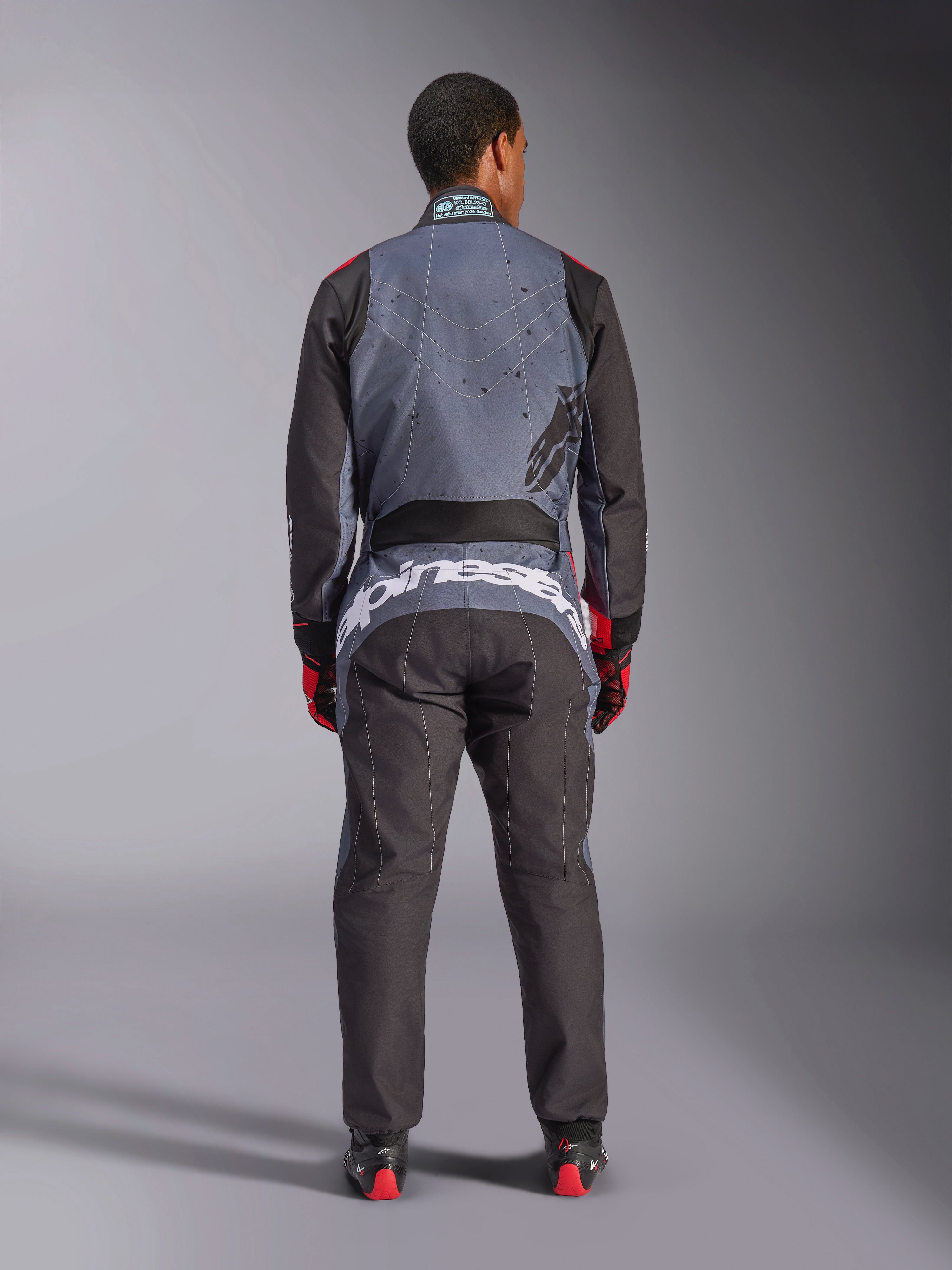 Alpinestars KMX-9 V3 Graphic 6 Suit, mono de karting, negro antracita y rojo, vista trasera que muestra un gran logotipo blanco de Alpinestars en la parte baja de la espalda, parte superior del cuerpo gris con gráficos de salpicaduras negras, brazos y piernas negros, usado con guantes y botas de carreras en rojo y negro.