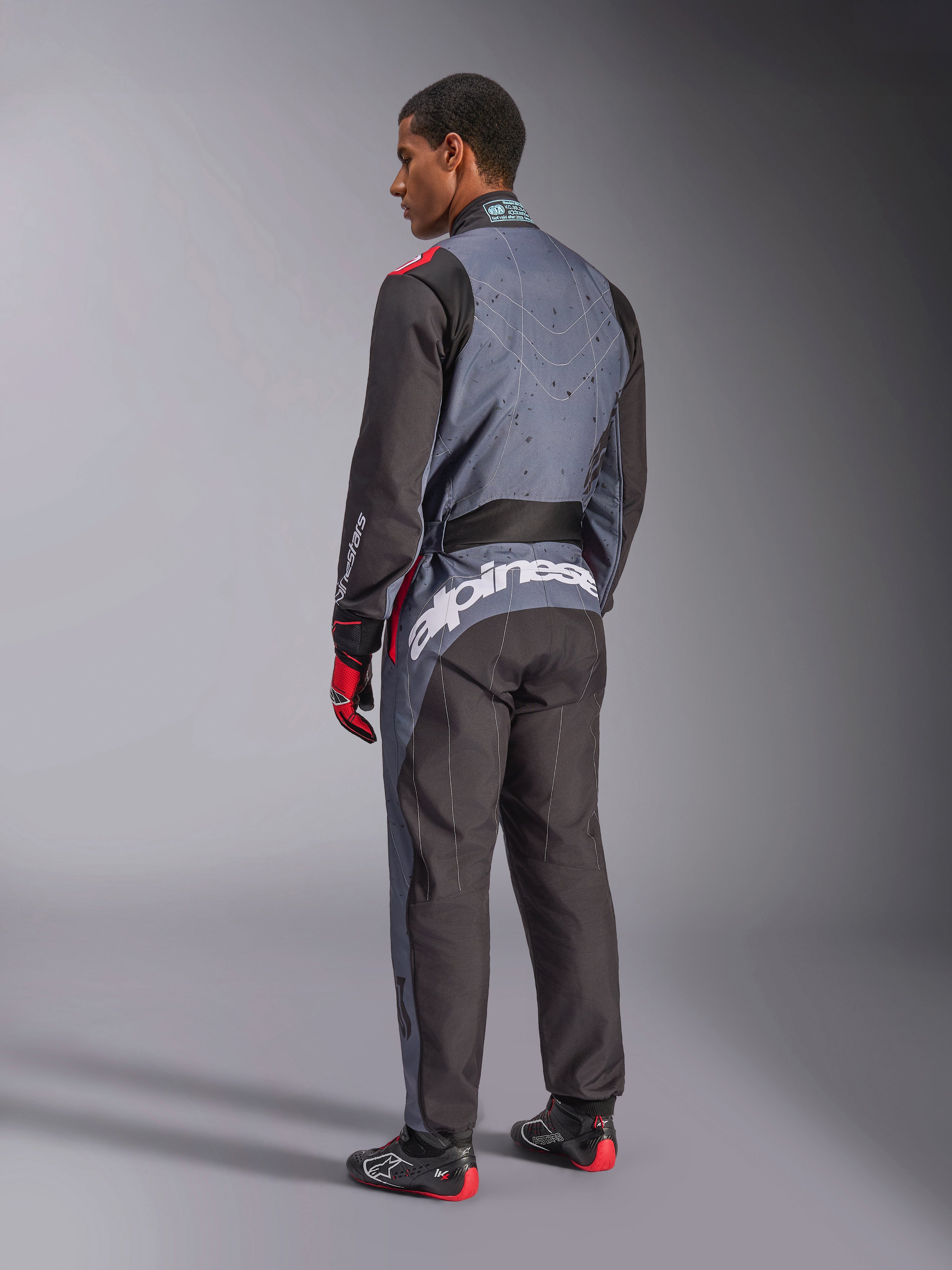 Vista posterior de un modelo con el mono Alpinestars KMX-9 V3 Graphic 6, traje de karting, Antracita Negro Rojo, con la marca Alpinestars en blanco destacada en la parte inferior de la espalda, diseño de paneles negros y grises con detalles rojos, usado con guantes y calzado de competición a juego.