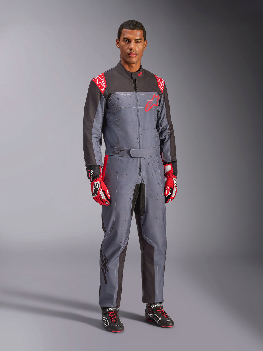 Mono de karting Alpinestars KMX-9 V3 Graphic 6, gris y antracita con acentos rojos, con un gráfico de salpicaduras único en el pecho y las piernas, combinado con guantes de carreras rojos y botas de karting negras, fotografía de estudio de cuerpo completo.
