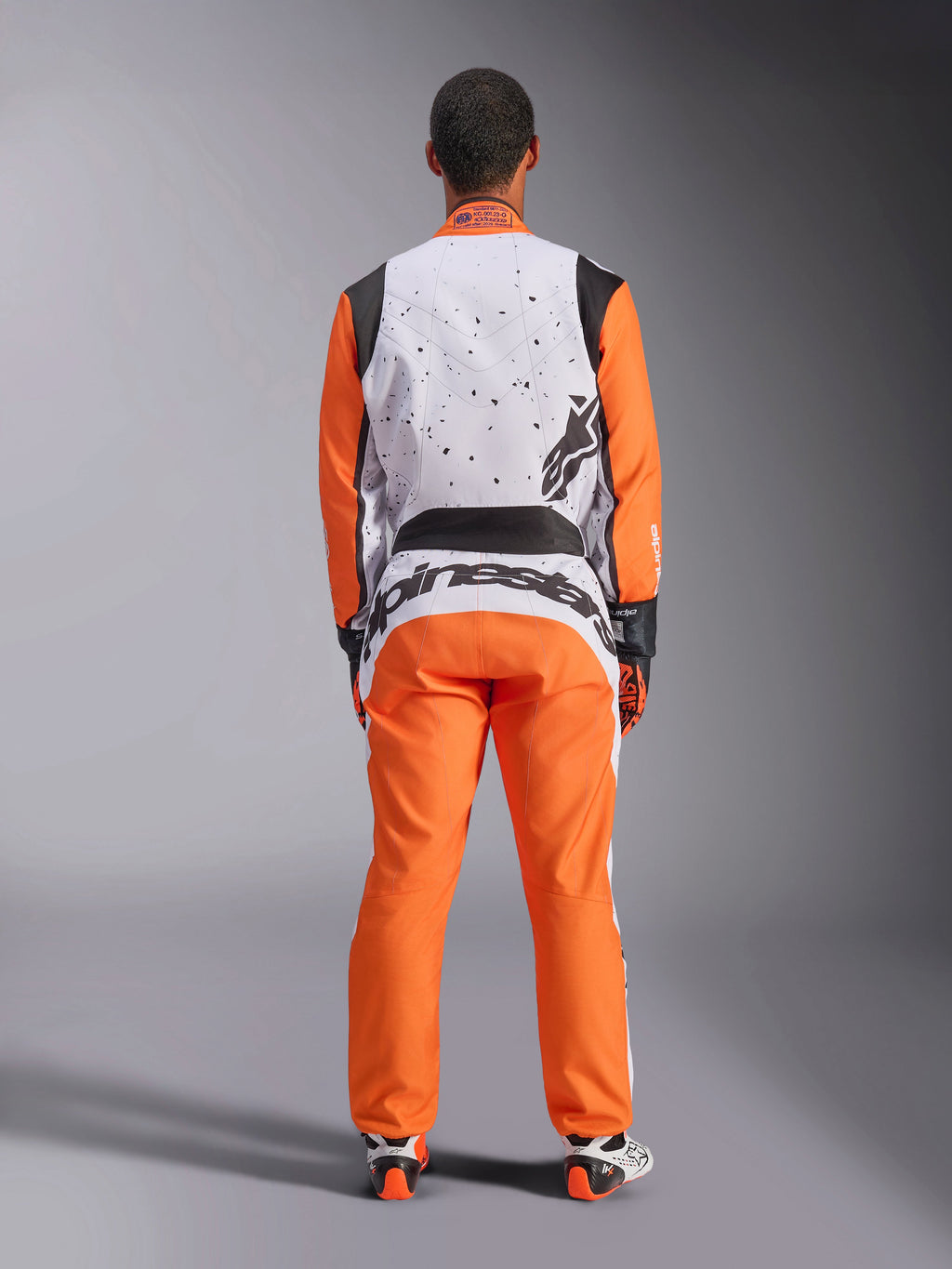 Vista posterior de un piloto con el mono Alpinestars KMX-9 V3 Graphic 6 en Blanco Naranja Negro, parte superior de la espalda blanca con gráficos de motas negras, mangas y pantalones naranjas, logotipo grande de Alpinestars en negro en la parte inferior de la espalda, coordinado con guantes y botas de carreras para karting.