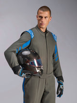 Piloto con el traje Alpinestars GP Race V3 Suit FIA en Anthracite Blue, sujetando un casco de carreras de fibra de carbono con visera iridiscente, equipamiento profesional de automovilismo con detalles azules en los hombros y guantes de carreras a juego.