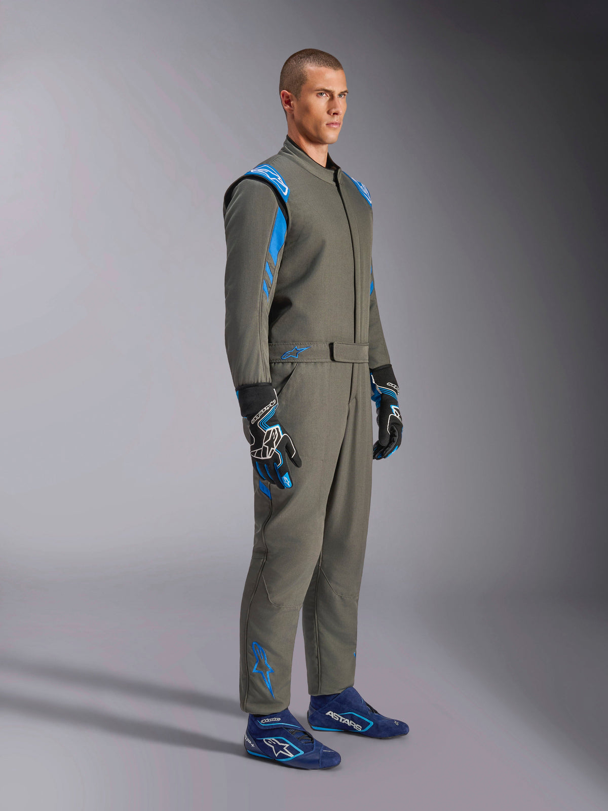 Alpinestars GP Race V3 Suit FIA en Anthracite Blue, mono de automovilismo de una pieza usado por un modelo masculino, con detalles en azul brillante en hombros y tobillos, combinado con guantes de carreras y calzado de competición azul, equipamiento profesional de automovilismo.
