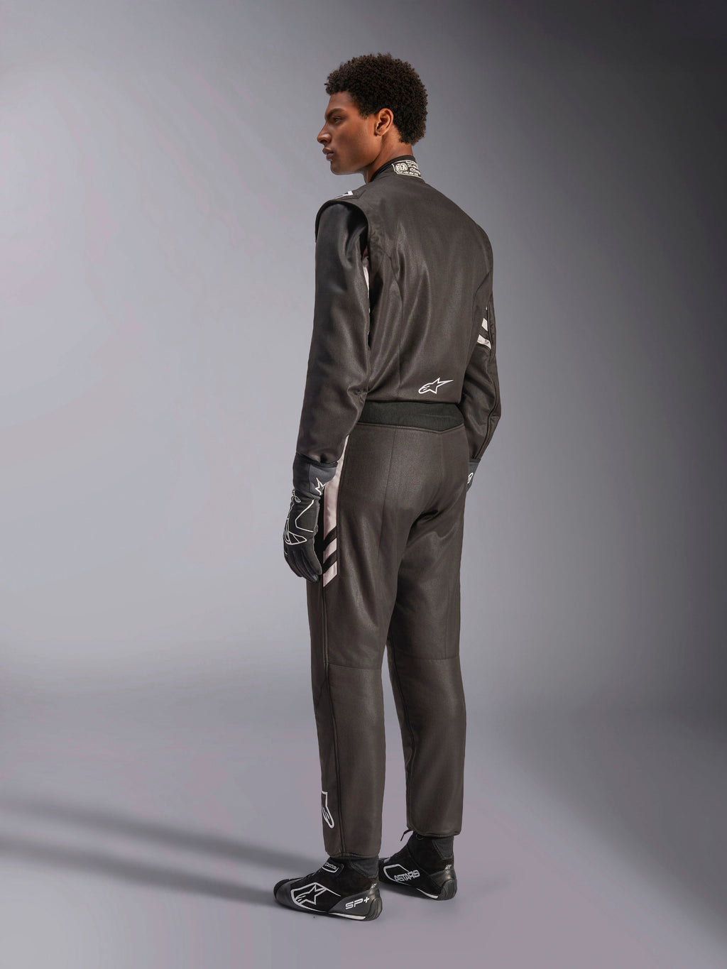 Alpinestars GP Race V3 Suit FIA, mono de automovilismo negro con detalles plateados, visto desde una perspectiva trasera de tres cuartos en un modelo, combinado con guantes y botas de carreras negros a juego, equipamiento profesional de automovilismo con un diseño de dos capas.