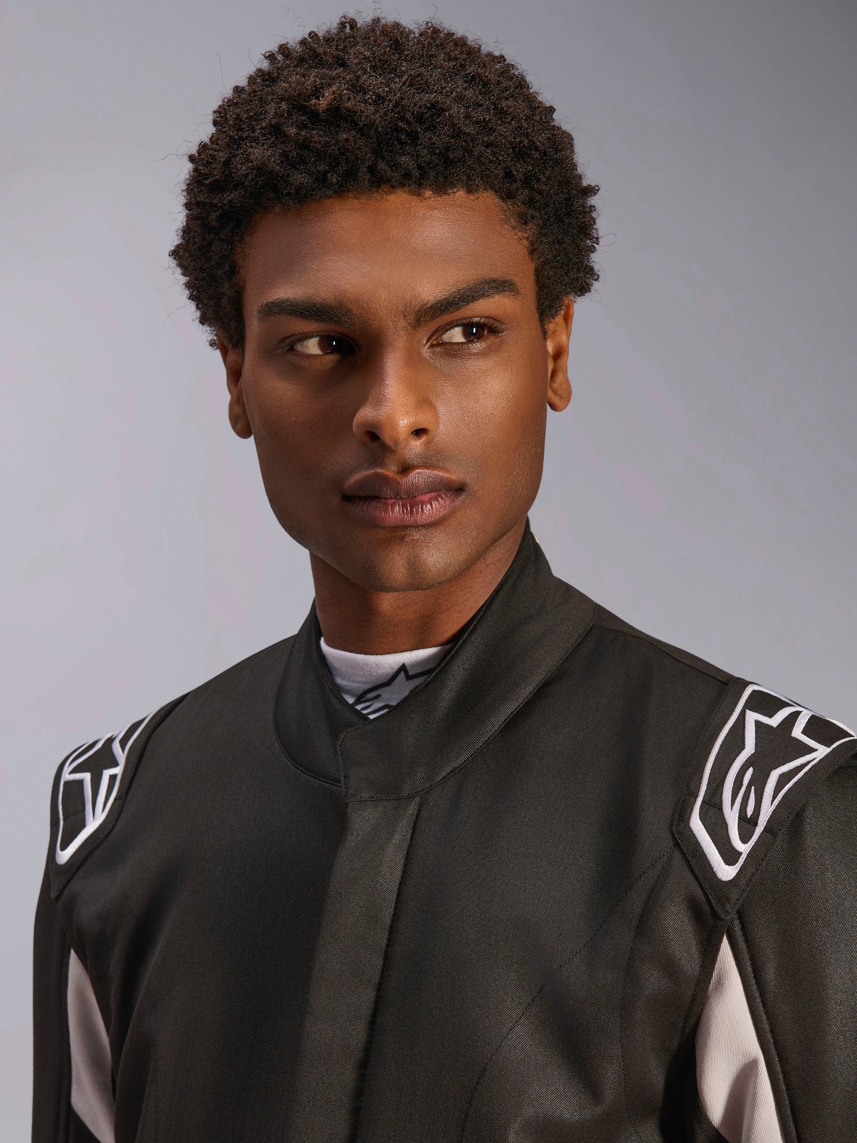 Alpinestars GP Race V3 Suit FIA, mono de automovilismo negro usado por un hombre, vista en primer plano que muestra el cuello suave y los logotipos de estrellas blancas en las hombreras, equipo de protección profesional para automovilismo.