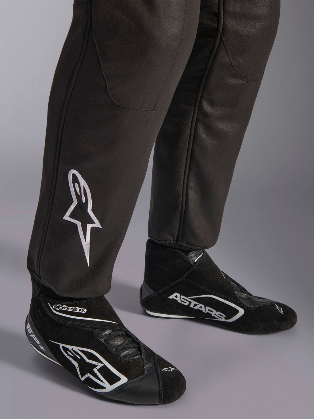 Alpinestars GP Race V3 Suit FIA, primer plano de la parte inferior de las piernas y botas de carreras, negro con detalles plateados, con el logotipo de la estrella bordado en blanco en la espinilla y puños acanalados, equipo de protección profesional para automovilismo.