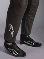 Alpinestars GP Race V3 Suit FIA, primer plano de la parte inferior de las piernas y botas de carreras, negro con detalles plateados, con el logotipo de la estrella bordado en blanco en la espinilla y puños acanalados, equipo de protección profesional para automovilismo.