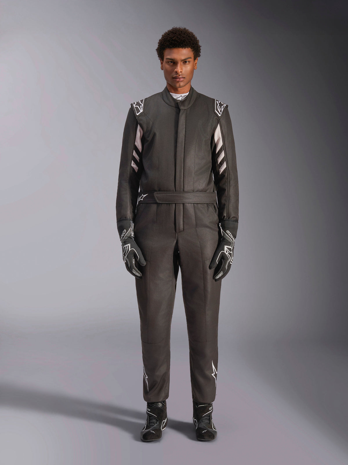 Alpinestars GP Race V3 Suit FIA, mono de automovilismo, negro y plata, vista frontal del piloto con equipamiento de carreras completo incluyendo guantes y botas, diseño profesional con logotipos de Alpinestars en hombros y cinturón
