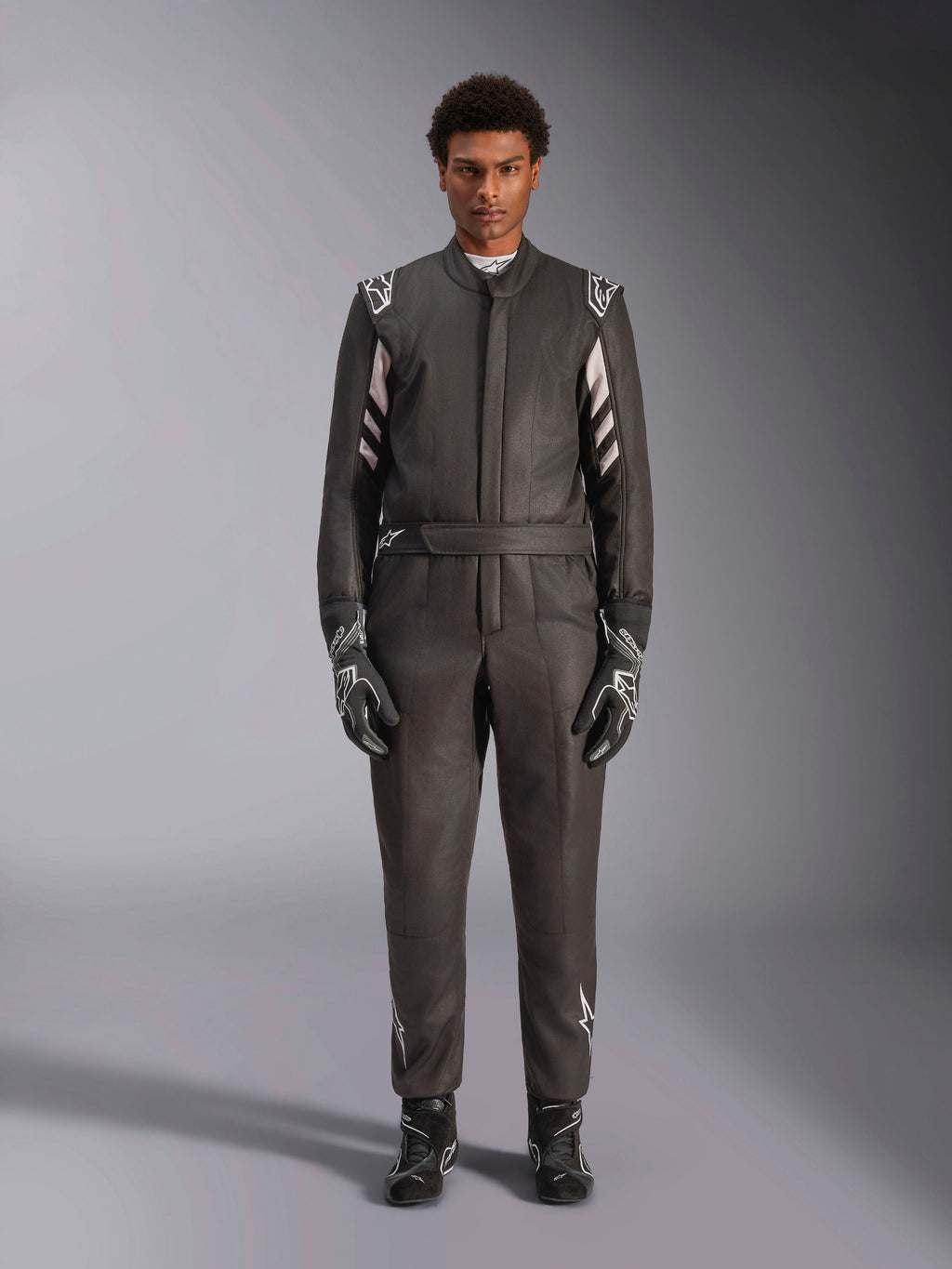 Alpinestars GP Race V3 Suit FIA, mono de automovilismo, negro y plata, vista frontal del piloto con equipamiento de carreras completo incluyendo guantes y botas, diseño profesional con logotipos de Alpinestars en hombros y cinturón