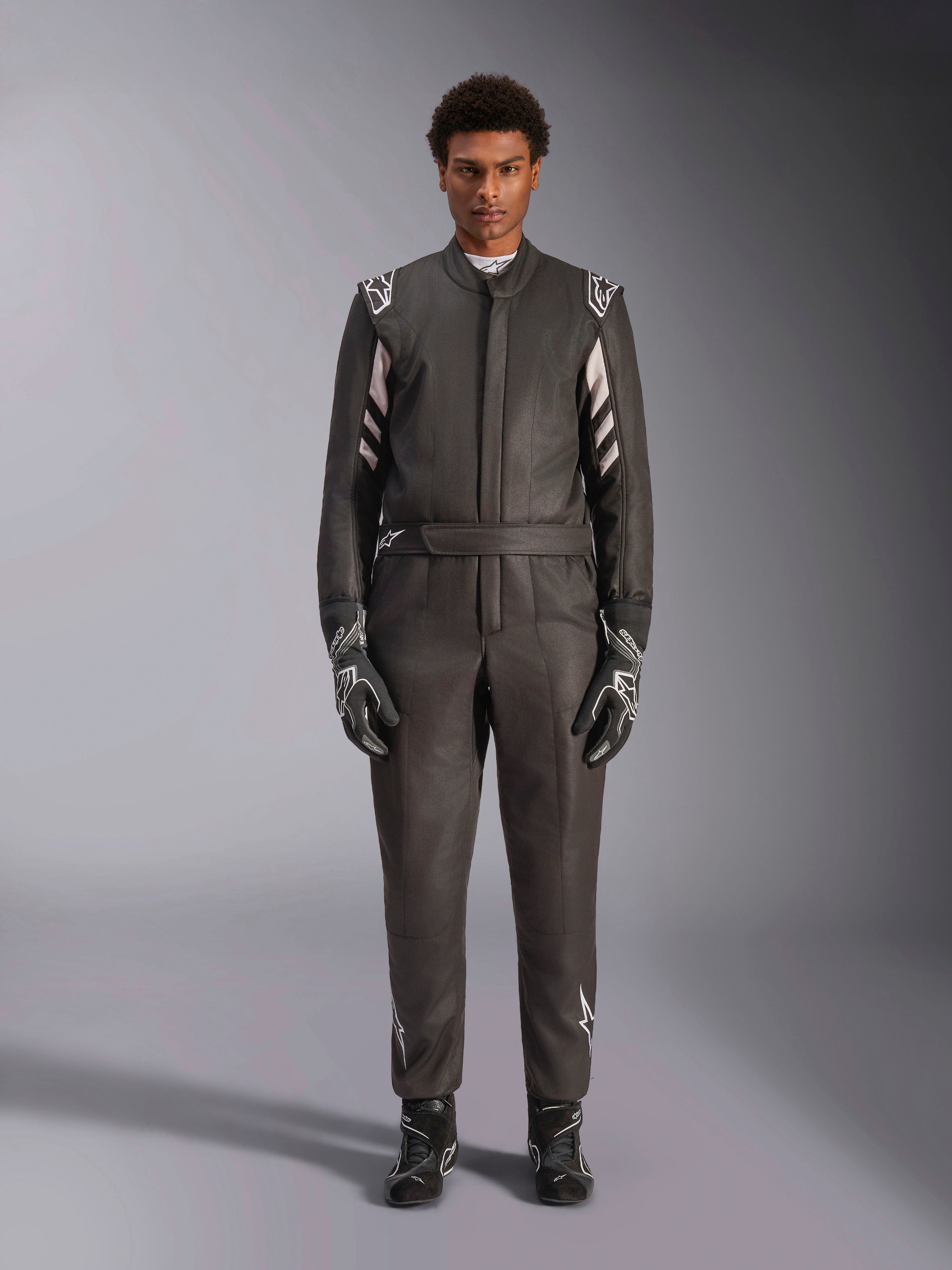 Alpinestars GP Race V3 Suit FIA, mono de automovilismo, negro y plata, vista frontal del piloto con equipamiento de carreras completo incluyendo guantes y botas, diseño profesional con logotipos de Alpinestars en hombros y cinturón