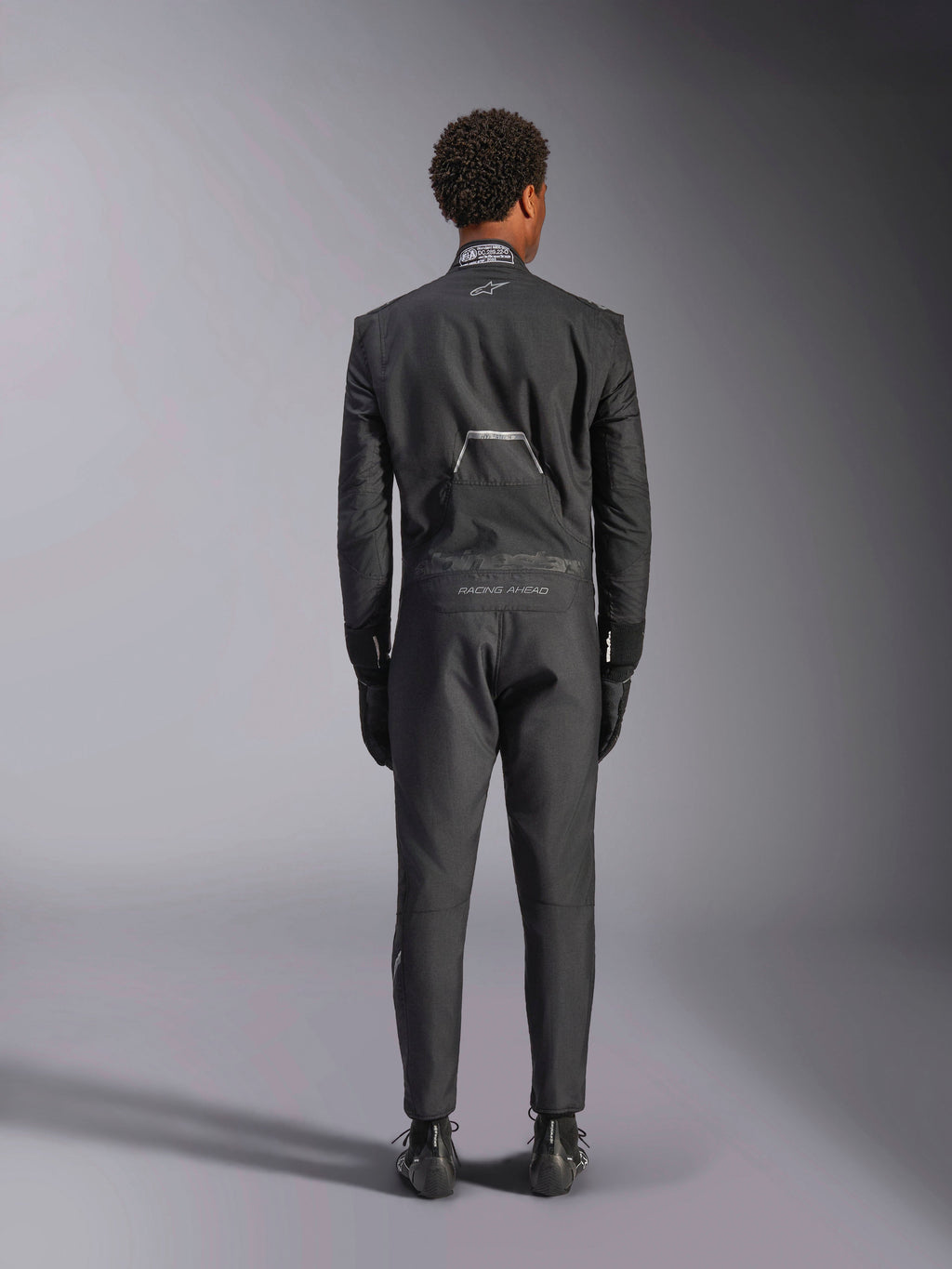 Mono Alpinestars Hypertech V3, Black Dark Shadow, vista posterior de un mono de automovilismo profesional de una pieza, con etiqueta de certificación FIA en el cuello y logotipos 'Alpinestars' y 'RACING AHEAD' en la parte inferior de la espalda, usado con guantes y botas de competición negros a juego.