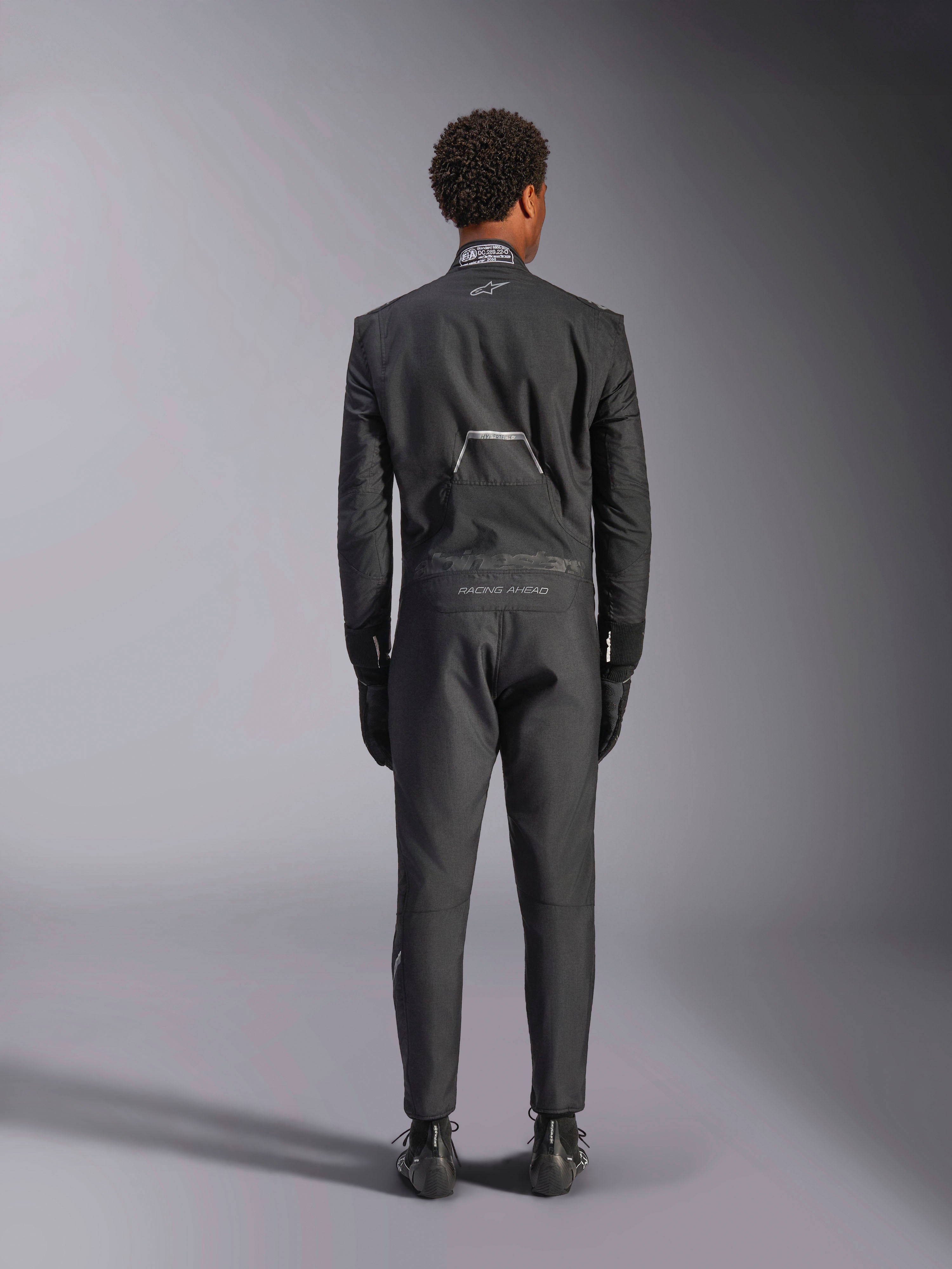 Mono Alpinestars Hypertech V3, Black Dark Shadow, vista posterior de un mono de automovilismo profesional de una pieza, con etiqueta de certificación FIA en el cuello y logotipos 'Alpinestars' y 'RACING AHEAD' en la parte inferior de la espalda, usado con guantes y botas de competición negros a juego.
