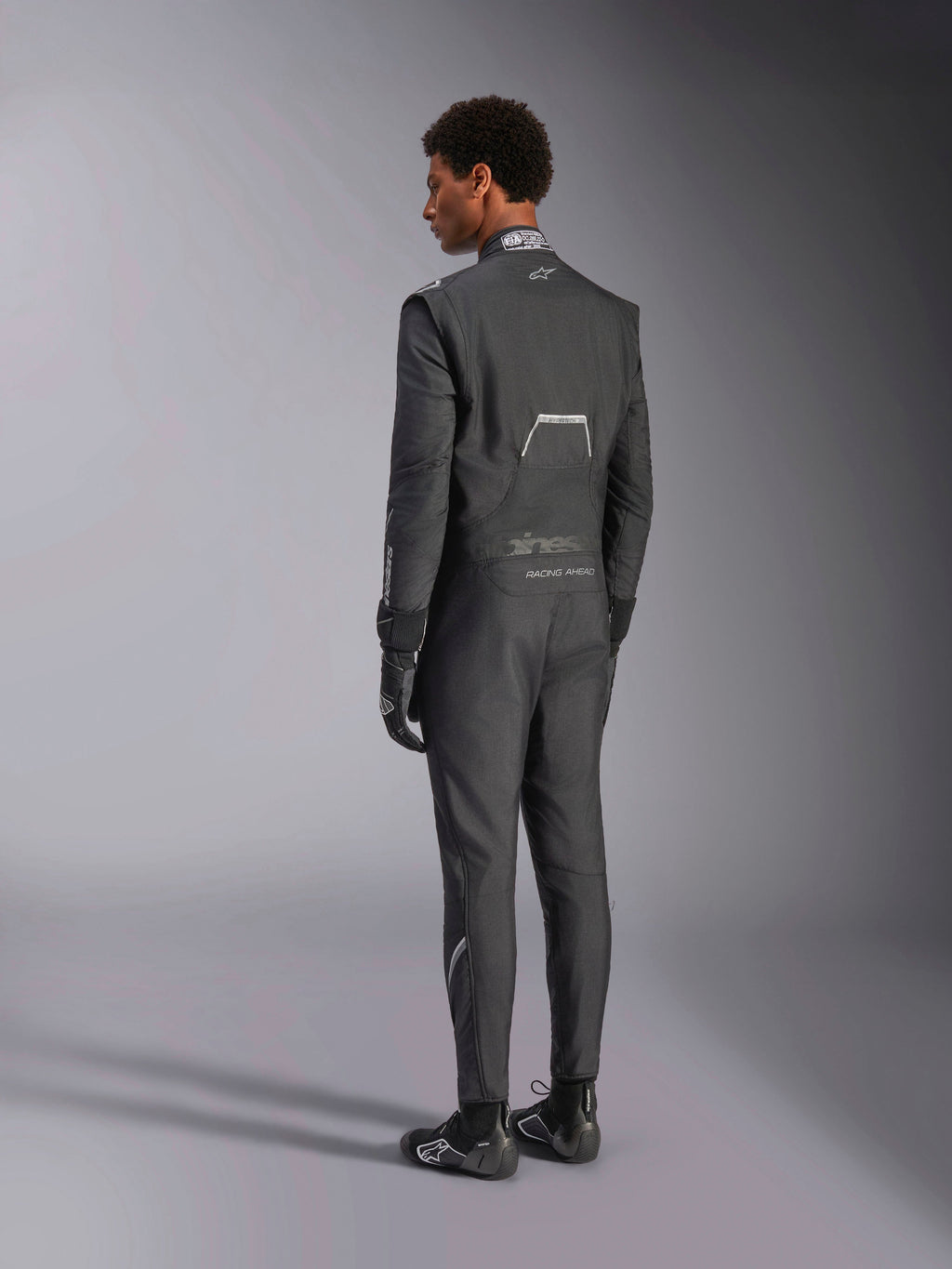 Mono Alpinestars Hypertech V3, Negro y Dark Shadow, vista trasera de modelo masculino con traje de carreras profesional, guantes y botas negros a juego, confeccionado en Nomex ligero con certificación FIA en el cuello, ajuste atlético para automovilismo.