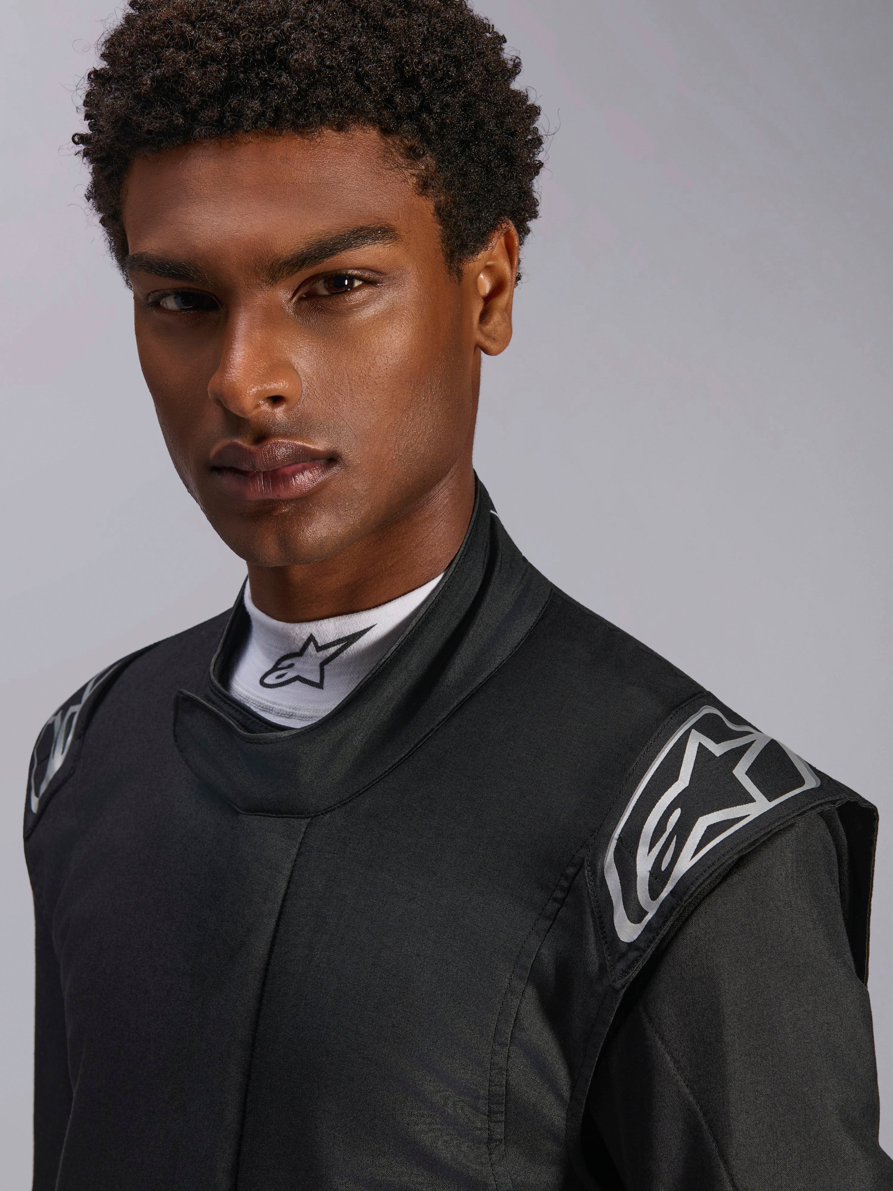 Mono Alpinestars Hypertech V3, traje de automovilismo Black Dark Shadow, primer plano de modelo masculino, logotipos plateados en los hombros, cuello alto con camiseta blanca Alpinestars de cuello falso, equipo de protección profesional para automovilismo