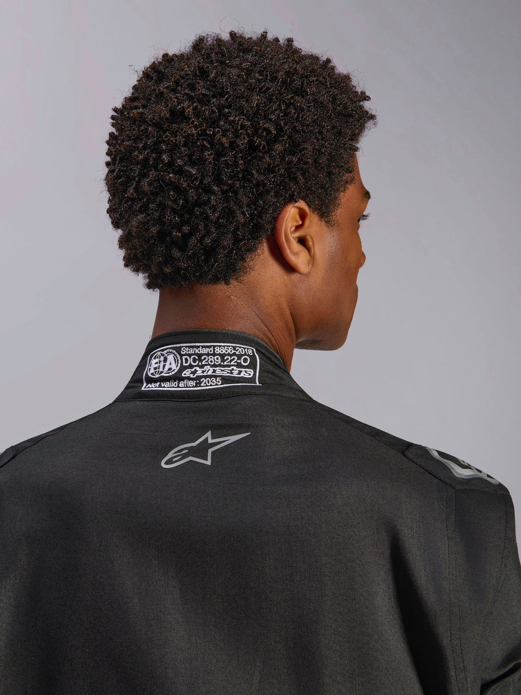 Mono Alpinestars Hypertech V3, Black Dark Shadow, primer plano de la vista trasera del cuello con etiqueta de certificación FIA 8856-2018 y logotipo en la espalda, traje de carreras profesional ligero con corte atlético