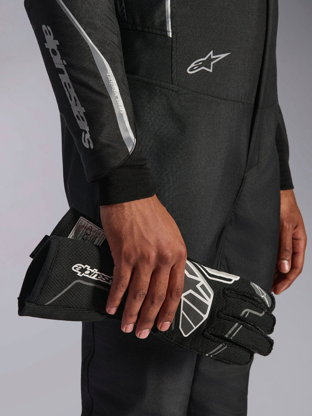 Mono Alpinestars Hypertech V3 en color Black Dark Shadow, detalle de primer plano de la manga y el torso con logotipos de Alpinestars en plateado, mano sosteniendo un guante de carreras negro con detalles blancos, equipamiento profesional de automovilismo