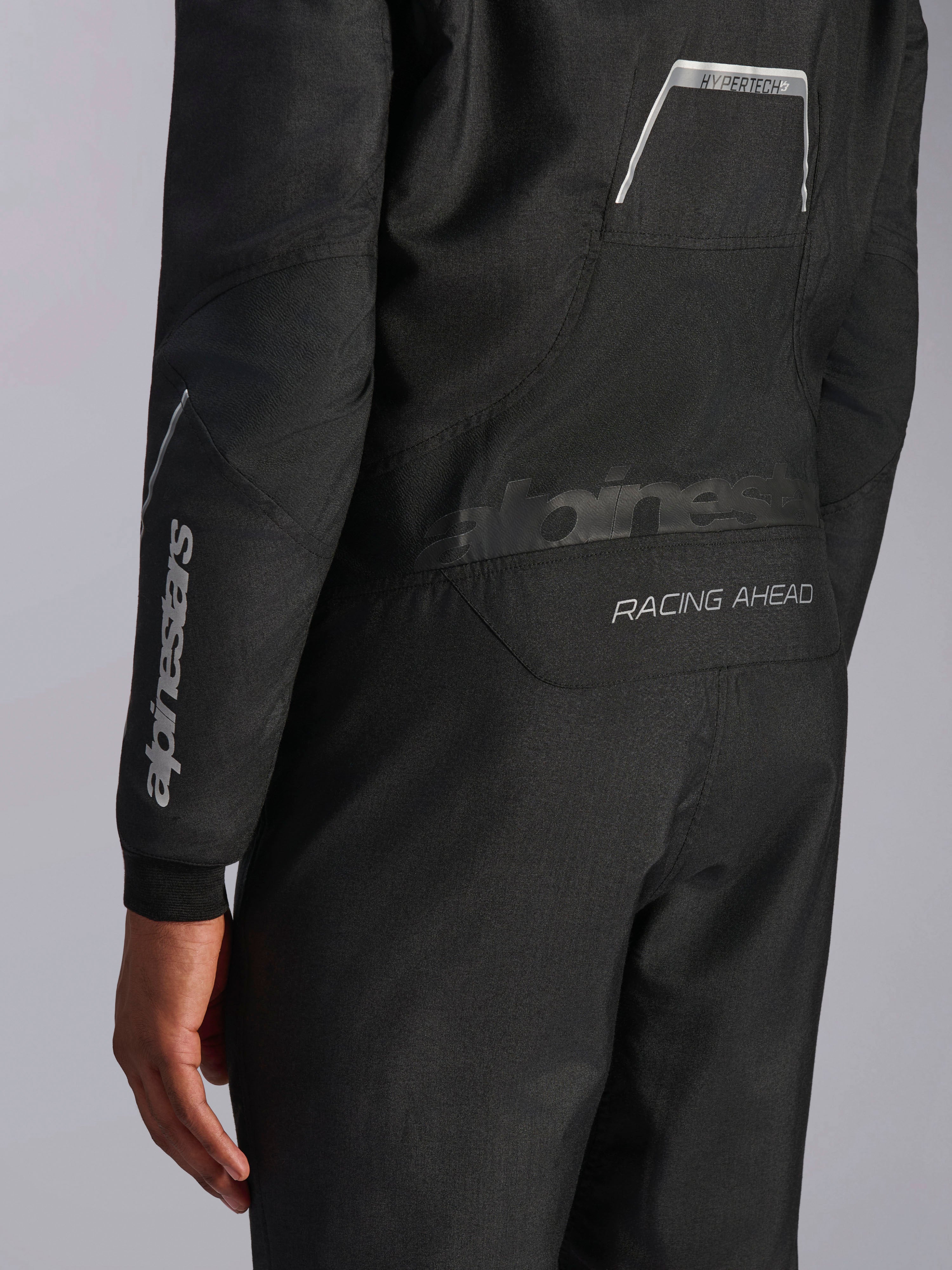 Primer plano del mono Alpinestars Hypertech V3 en Negro/Dark Shadow, que muestra los gráficos traseros 'RACING AHEAD' e 'HYPERTECH V3', con el logotipo blanco en la manga, equipamiento profesional para automovilismo.