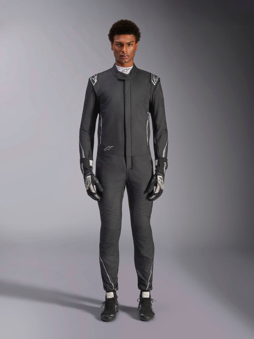Traje Alpinestars Hypertech V3, mono de automovilismo profesional, Negro y Dark Shadow, usado por un modelo con guantes y calzado de carreras a juego, corte atlético ajustado con detalles plateados y logotipos, equipamiento de alto rendimiento para automovilismo
