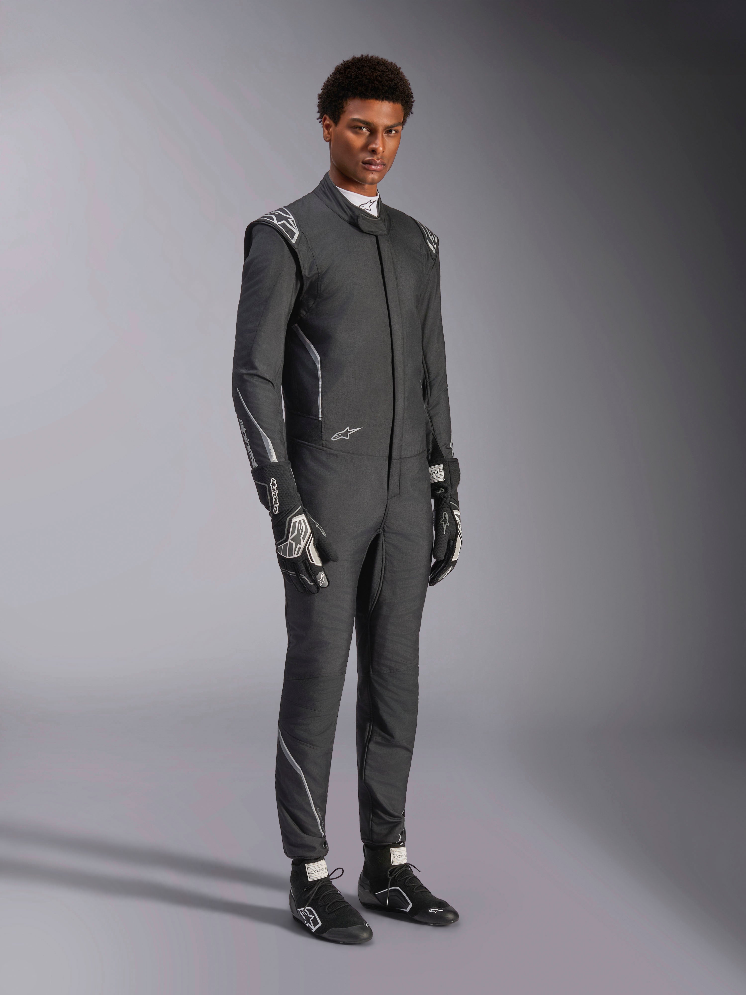 Mono Alpinestars Hypertech V3, traje de automovilismo, negro y Dark Shadow, usado por un modelo masculino con guantes y calzado de competición negros a juego, corte atlético ajustado, incluye logotipos de Alpinestars y detalles en gris claro, equipamiento de protección profesional para automovilismo.