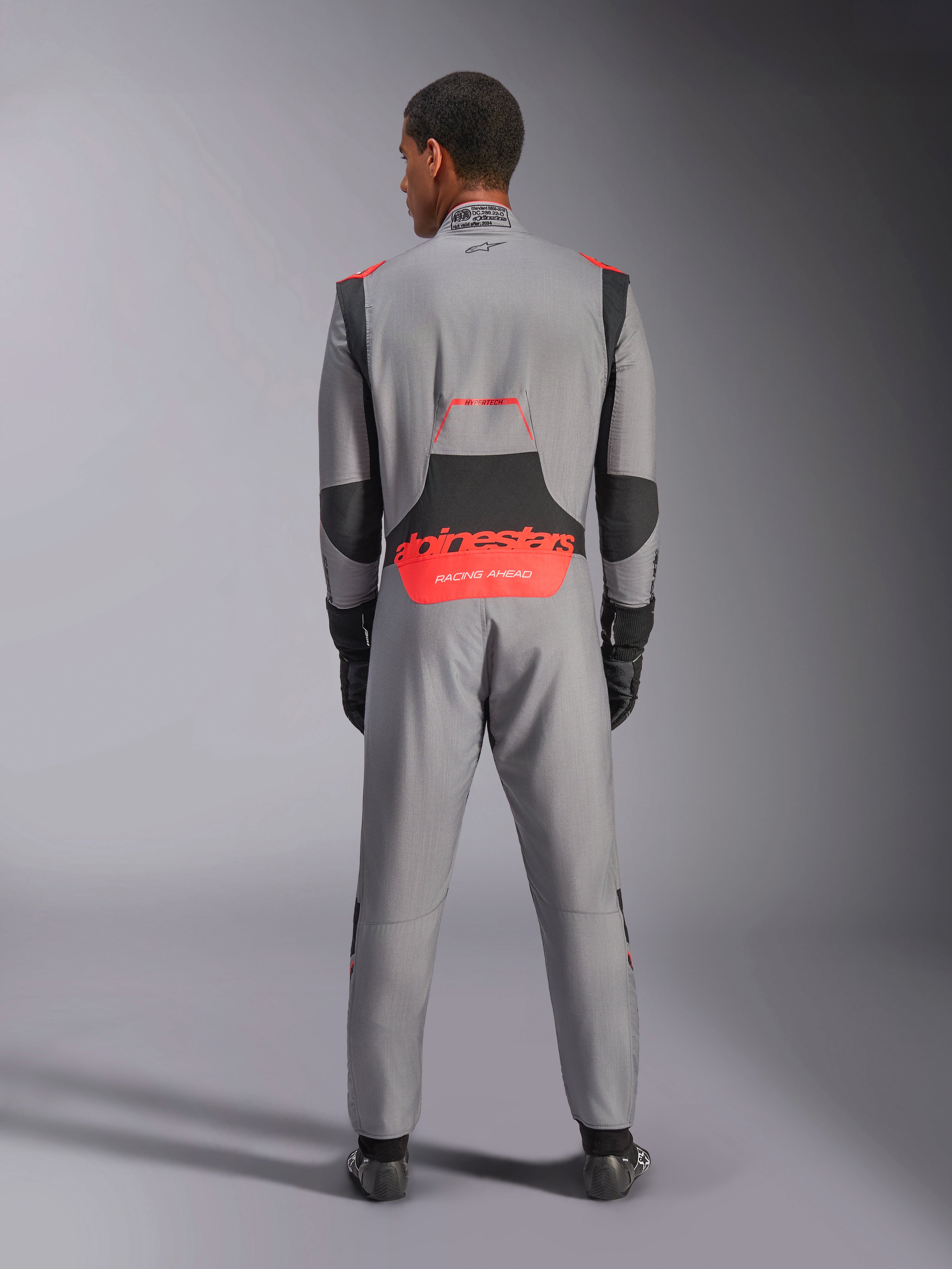 Mono Alpinestars Hypertech V3, traje de automovilismo profesional, gris medio con detalles en negro y rojo fluorescente, vista posterior en un modelo con el logotipo rojo de Alpinestars en la zona lumbar, confección ligera de Nomex para rendimiento en pista.