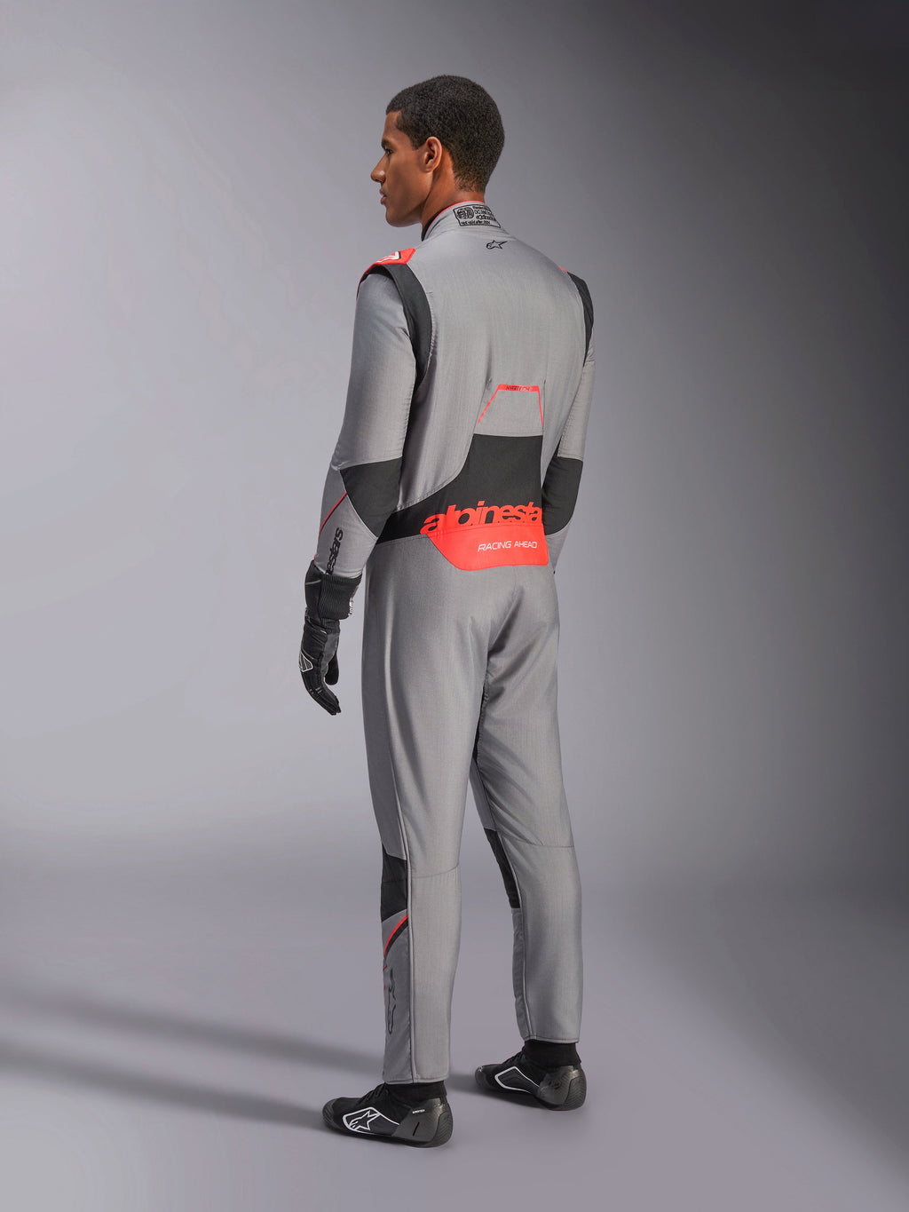 Mono Alpinestars Hypertech V3 en Gris Medio Negro Rojo Fluo, vista de tres cuartos trasera, con la marca Alpinestars en rojo llamativo en la parte inferior de la espalda, paneles elásticos negros y un ajuste atlético estilizado para automovilismo profesional.