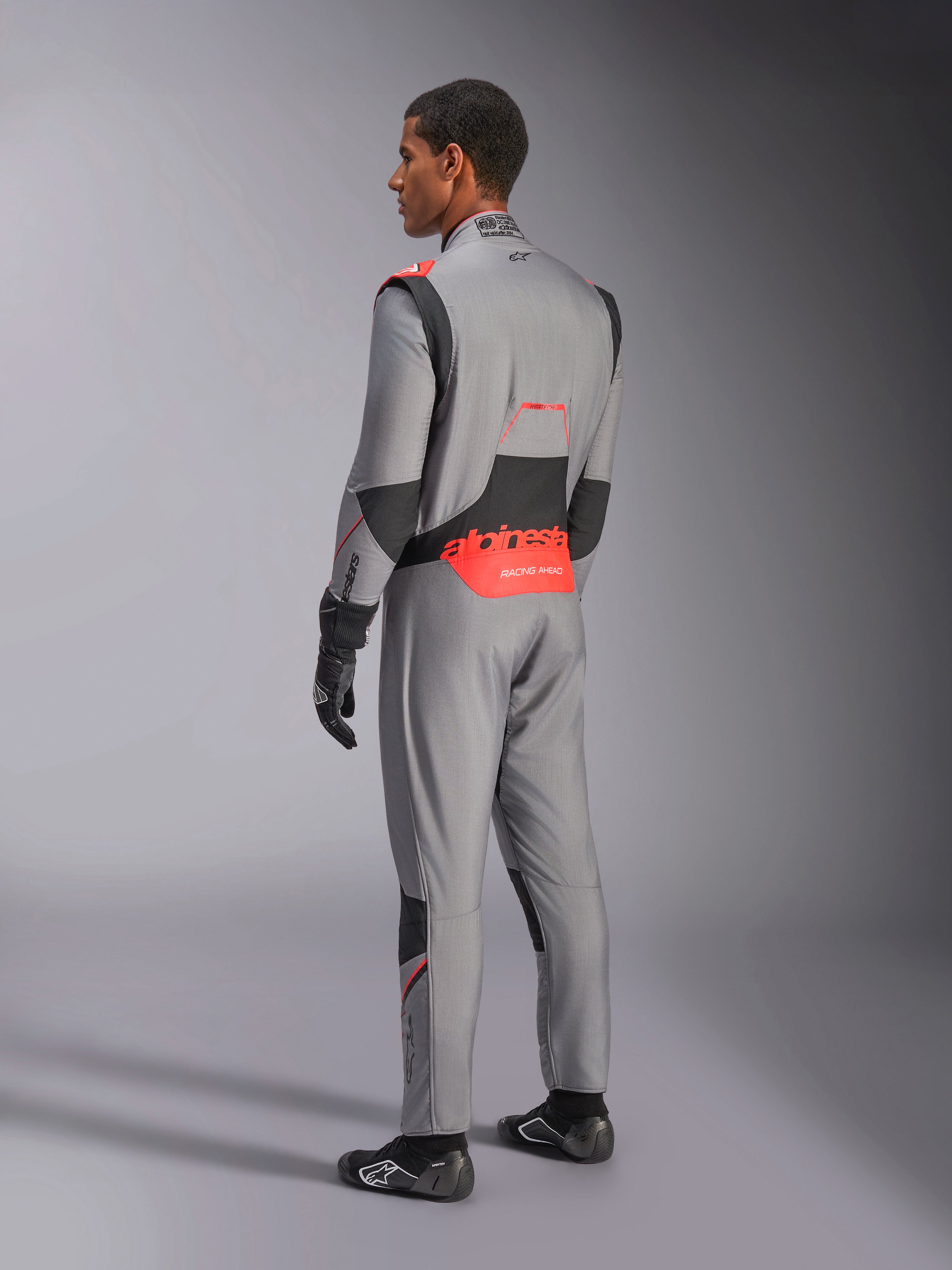 Mono Alpinestars Hypertech V3 en Gris Medio Negro Rojo Fluo, vista de tres cuartos trasera, con la marca Alpinestars en rojo llamativo en la parte inferior de la espalda, paneles elásticos negros y un ajuste atlético estilizado para automovilismo profesional.