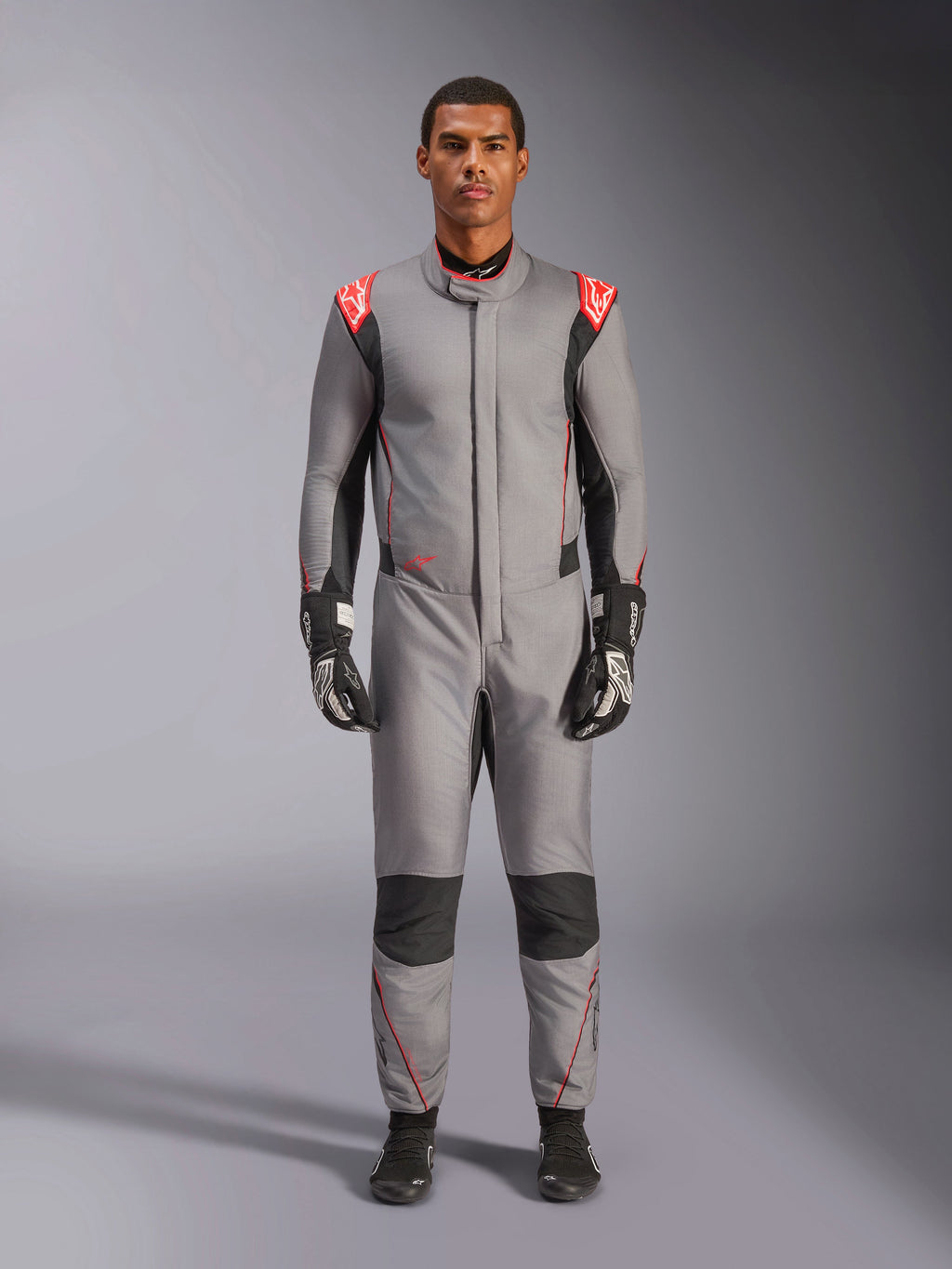 Mono Alpinestars Hypertech V3, traje de carreras profesional en gris medio, negro y rojo flúor, vestido por un modelo con guantes y calzado de competición negros, con un corte atlético ajustado, detalles en rojo flúor en los hombros y paneles negros en las rodillas y laterales.