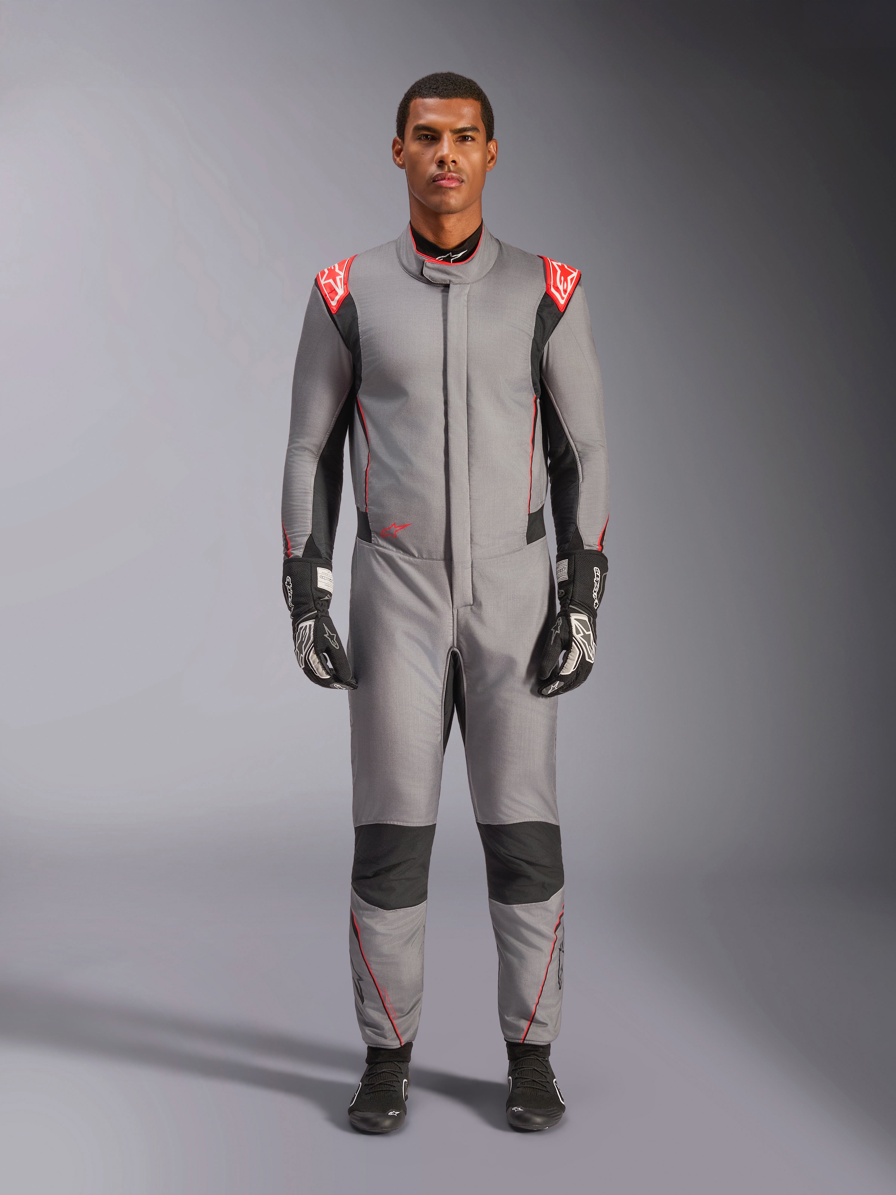 Mono Alpinestars Hypertech V3, traje de carreras profesional en gris medio, negro y rojo flúor, vestido por un modelo con guantes y calzado de competición negros, con un corte atlético ajustado, detalles en rojo flúor en los hombros y paneles negros en las rodillas y laterales.