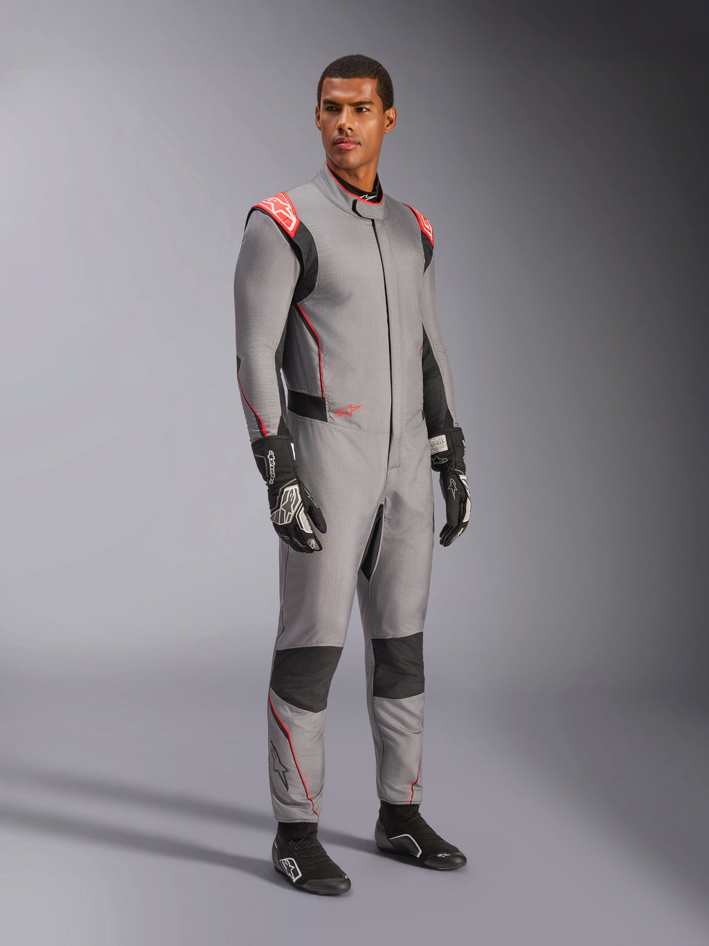 Modelo masculino con el mono Alpinestars Hypertech V3 en gris medio con acentos en negro y rojo flúor, traje de automovilismo profesional combinado con guantes y calzado de carreras negros, con confección ligera de dos capas de Nomex, detalles rojos en los hombros y un ajuste atlético
