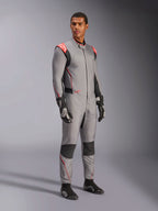 Modelo masculino con el mono Alpinestars Hypertech V3 en gris medio con acentos en negro y rojo flúor, traje de automovilismo profesional combinado con guantes y calzado de carreras negros, con confección ligera de dos capas de Nomex, detalles rojos en los hombros y un ajuste atlético