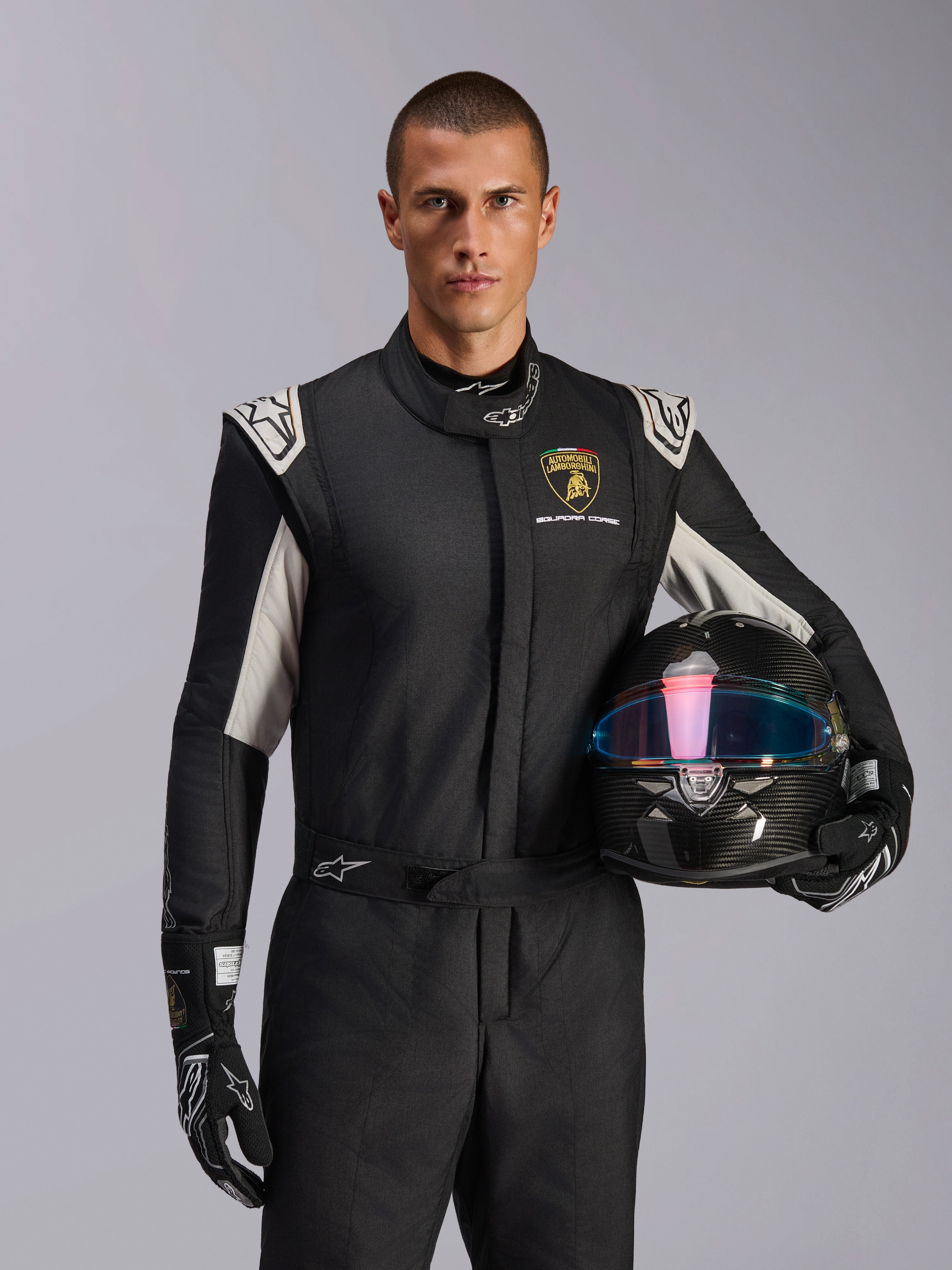 Male model wearing Limited Edition Lamborghini GP Tech V4 Suit, black racing suit with silver accents and Lamborghini Squadra Corse logo, holding a carbon fiber helmet, black racing gloves with white logos, professional auto racing gear