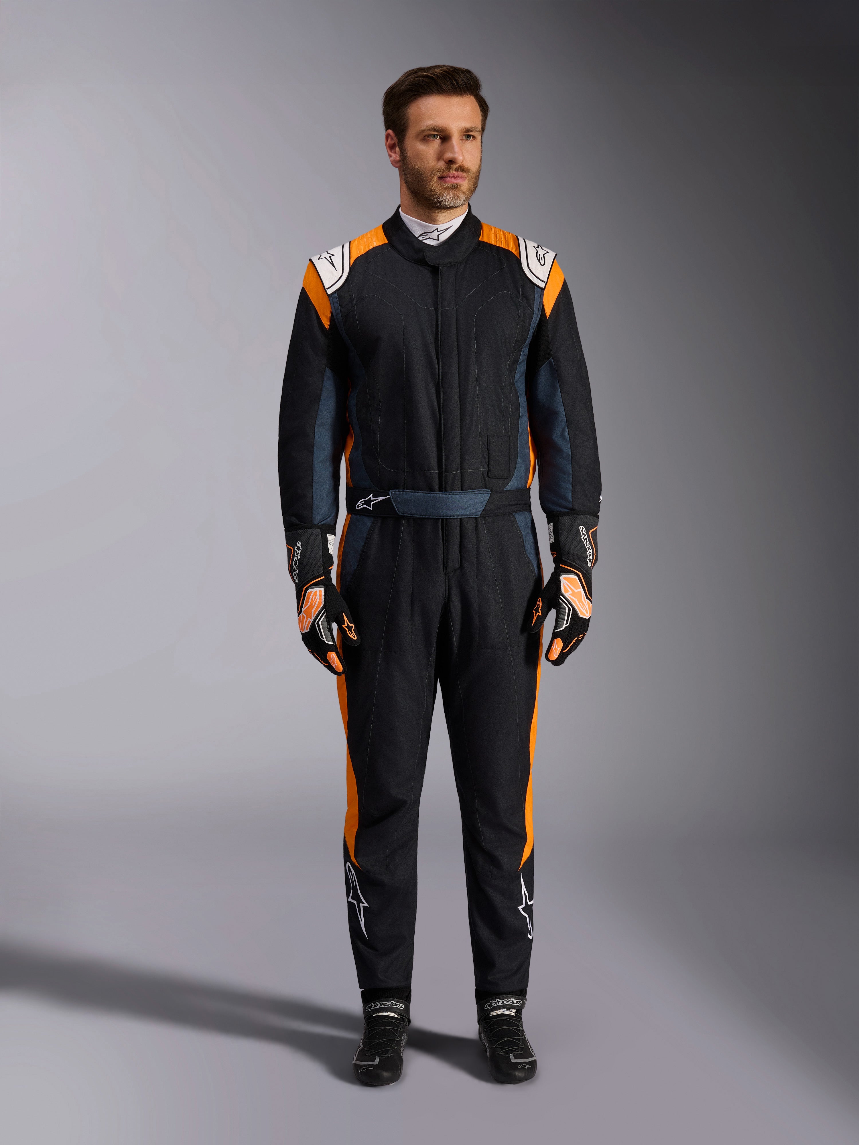 Alpinestars GP Pro Comp V2 FIA Suit, mono de carreras de automovilismo en negro asfalto y naranja flúor, modelo masculino con equipo de protección completo, guantes y calzado de carreras a juego, con logotipos blancos de Alpinestars y paneles laterales naranja vibrante, indumentaria profesional para automovilismo