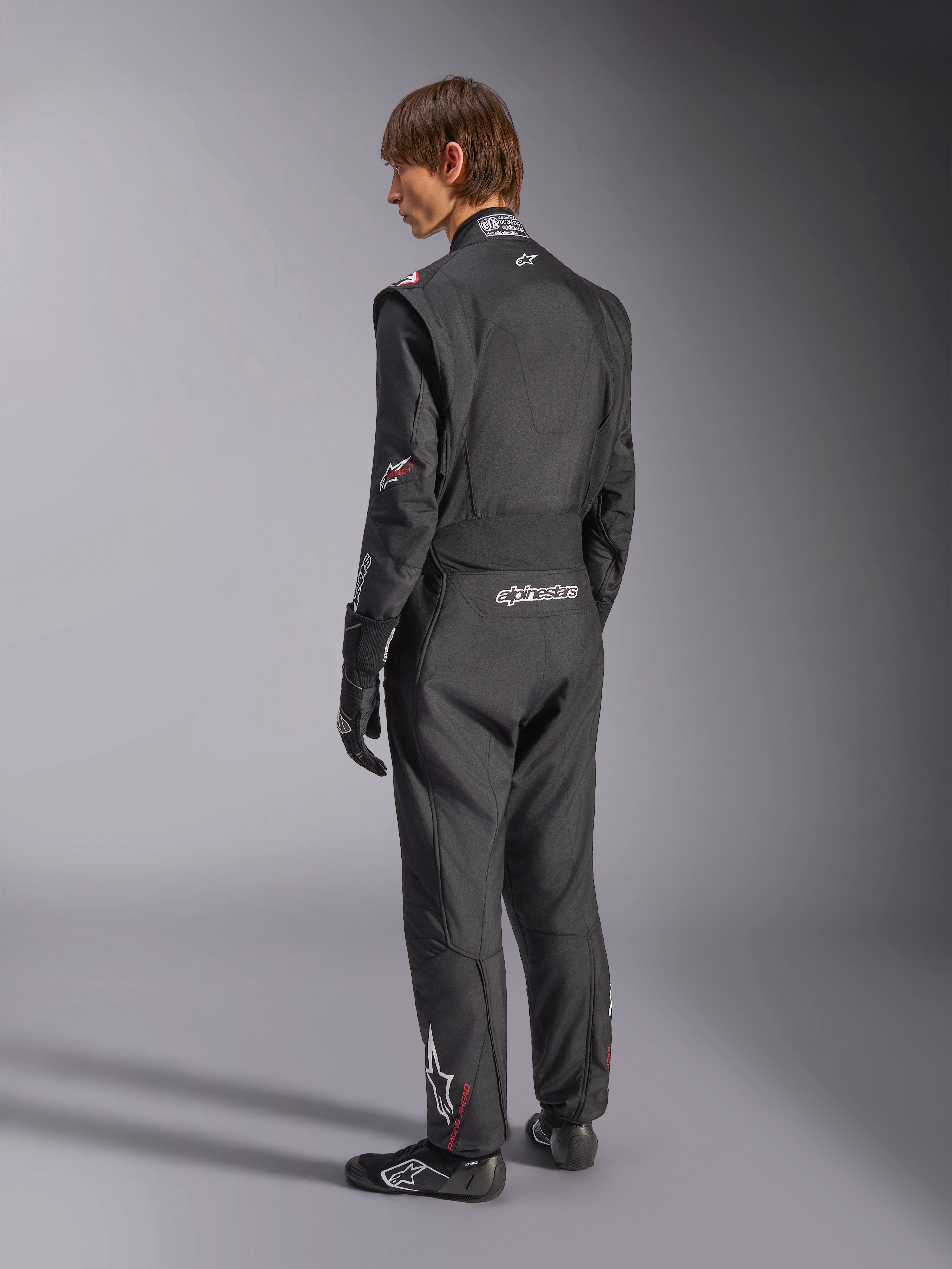 Alpinestars GP Tech V4 Suit, Auto Racing Suit, Black with white accents, back view of model wearing the suit with racing gloves and shoes, featuring white branding on the lower back and leg, professional motorsport safety gear