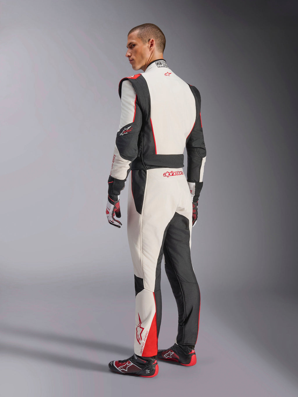 Mono Alpinestars GP Tech V4, traje de carreras en blanco, negro y rojo, vista trasera que muestra paneles elásticos ergonómicos en la zona lumbar y las piernas, combinado con guantes y calzado de competición a juego, equipamiento profesional para automovilismo.
