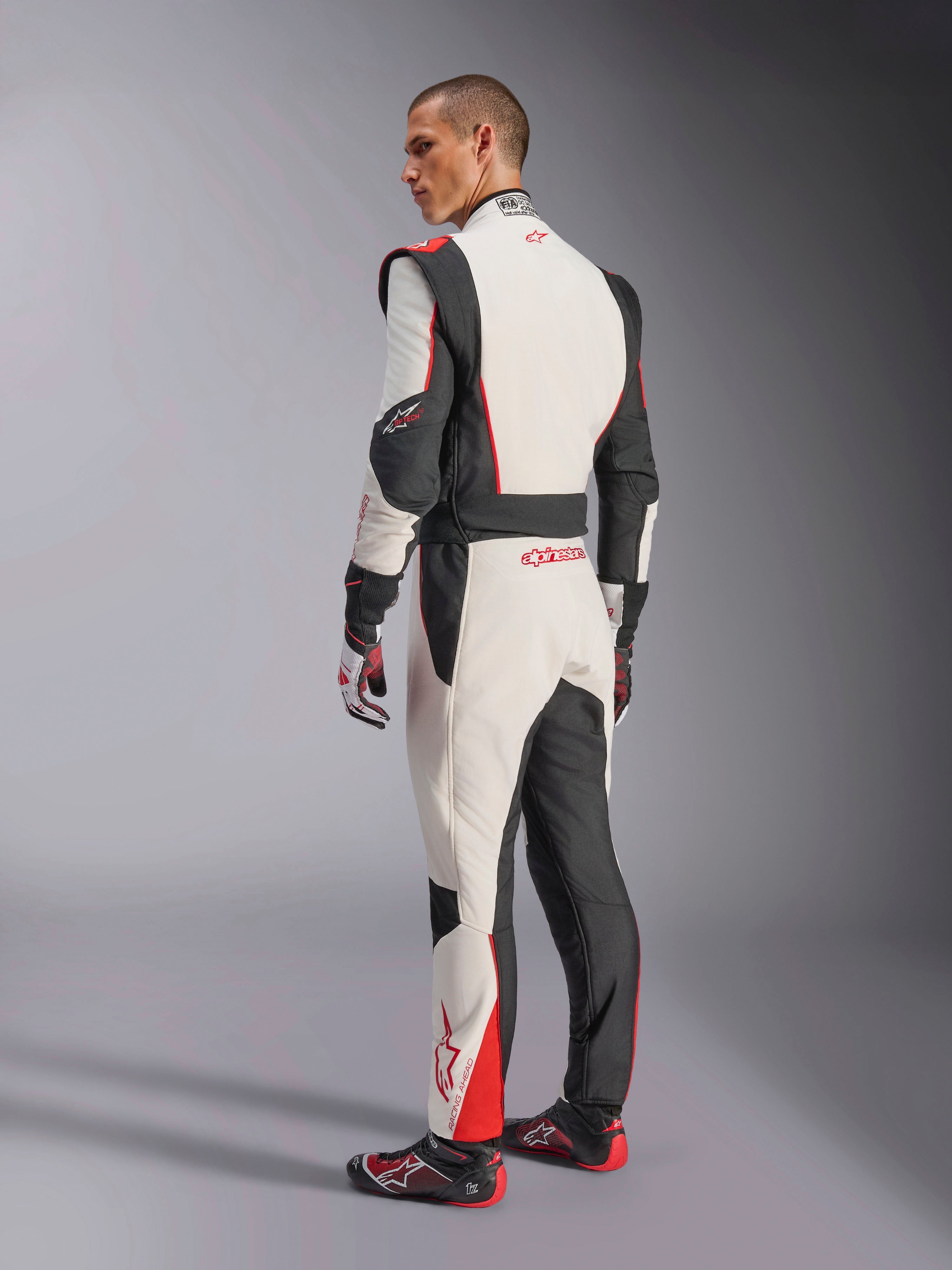 Mono Alpinestars GP Tech V4, traje de carreras en blanco, negro y rojo, vista trasera que muestra paneles elásticos ergonómicos en la zona lumbar y las piernas, combinado con guantes y calzado de competición a juego, equipamiento profesional para automovilismo.