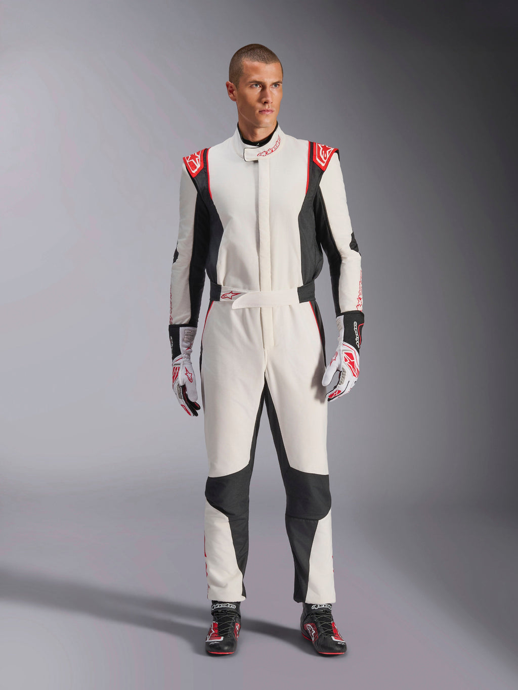 Alpinestars GP Tech V4 Suit, mono de automovilismo de una pieza en blanco, negro y rojo, usado por un modelo masculino con guantes y calzado de competición a juego, con logotipos rojos en los hombros y paneles laterales negros, equipamiento profesional de automovilismo para uso en pista.