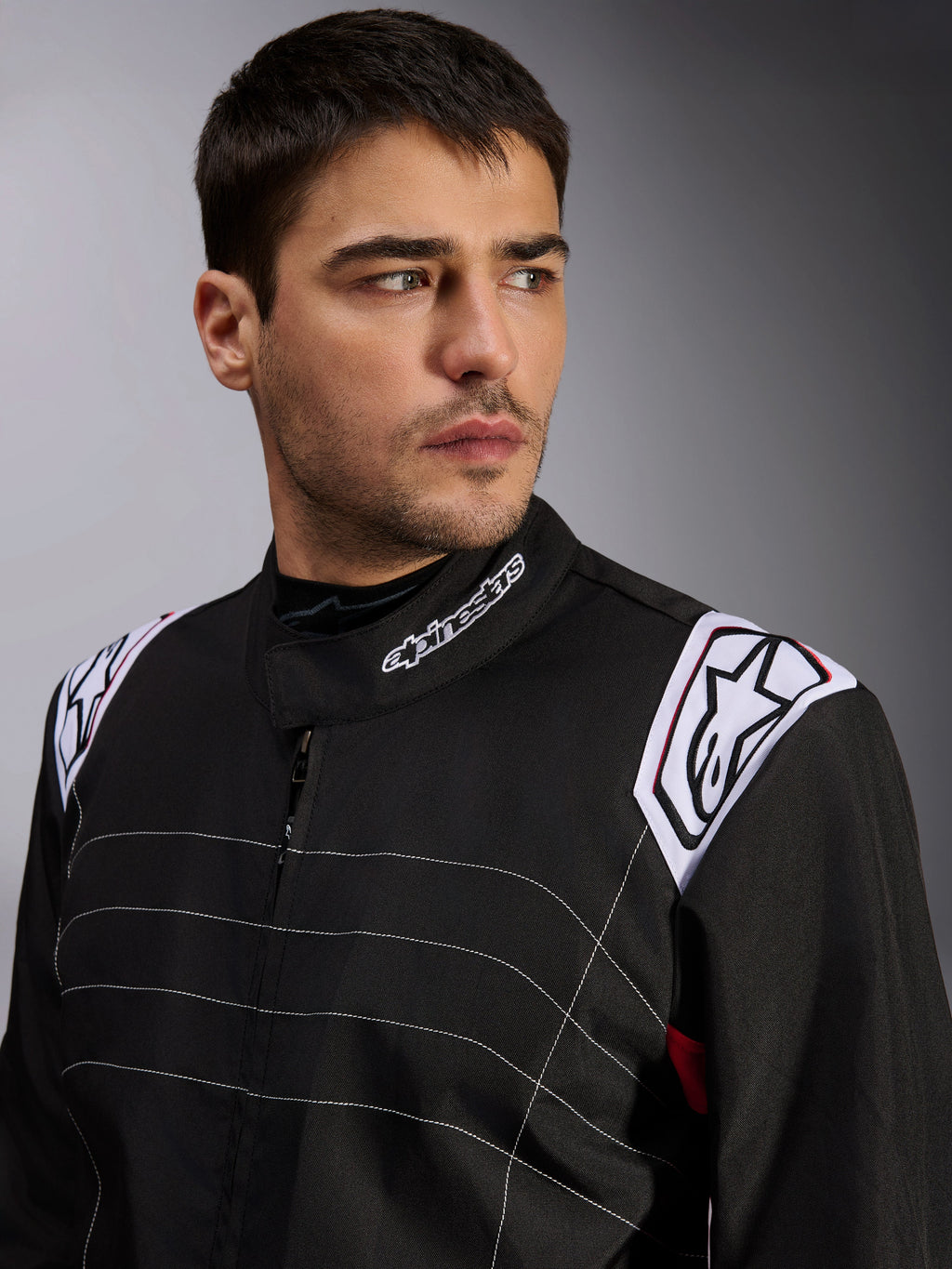 Mono Alpinestars KMX-9 V3, mono de karting negro llevado por un modelo masculino, primer plano del pecho y los hombros con logotipos blancos en el cuello y las hombreras, con costuras decorativas blancas y un diseño profesional para deportes de motor