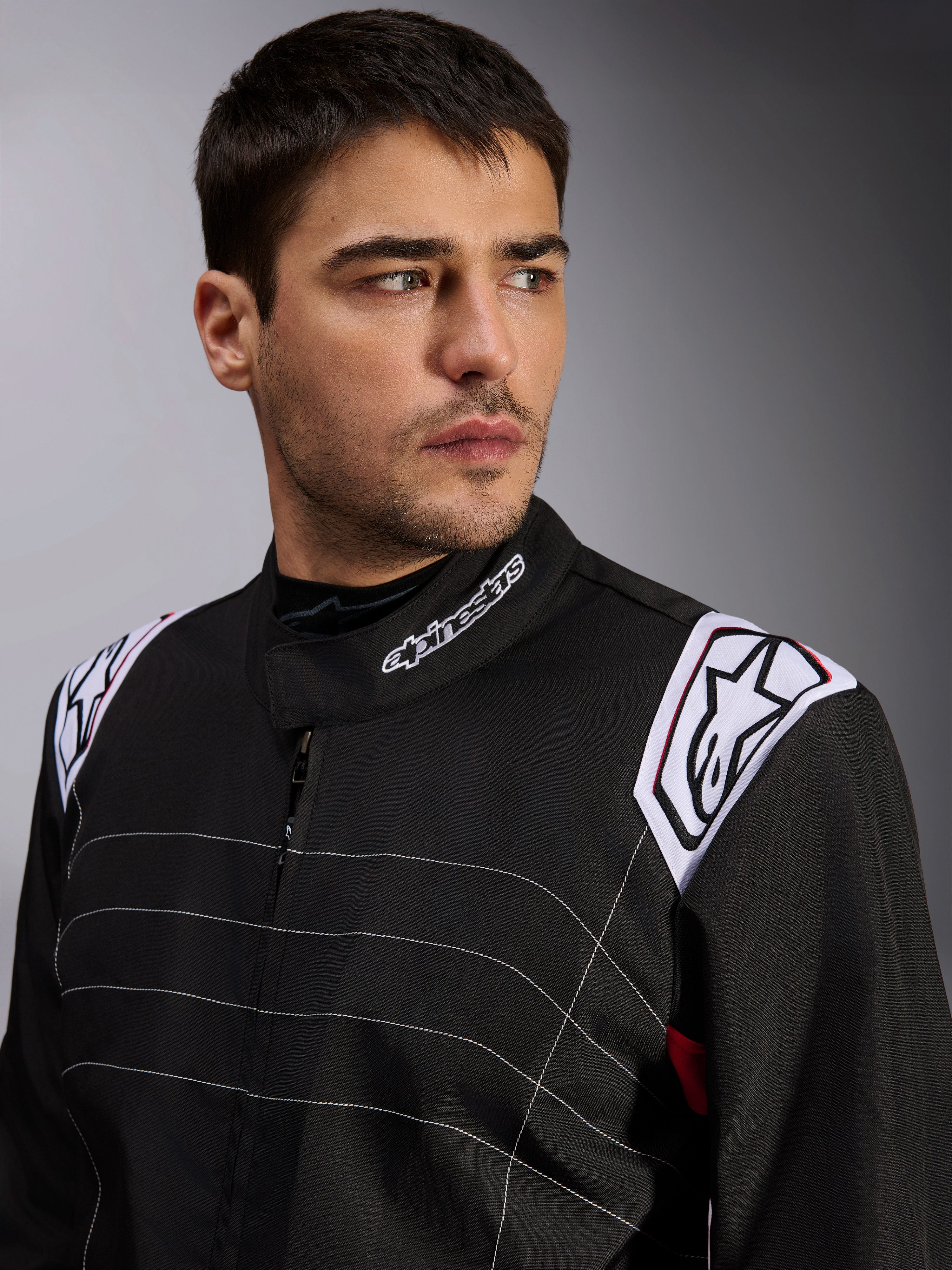 Mono Alpinestars KMX-9 V3, mono de karting negro llevado por un modelo masculino, primer plano del pecho y los hombros con logotipos blancos en el cuello y las hombreras, con costuras decorativas blancas y un diseño profesional para deportes de motor