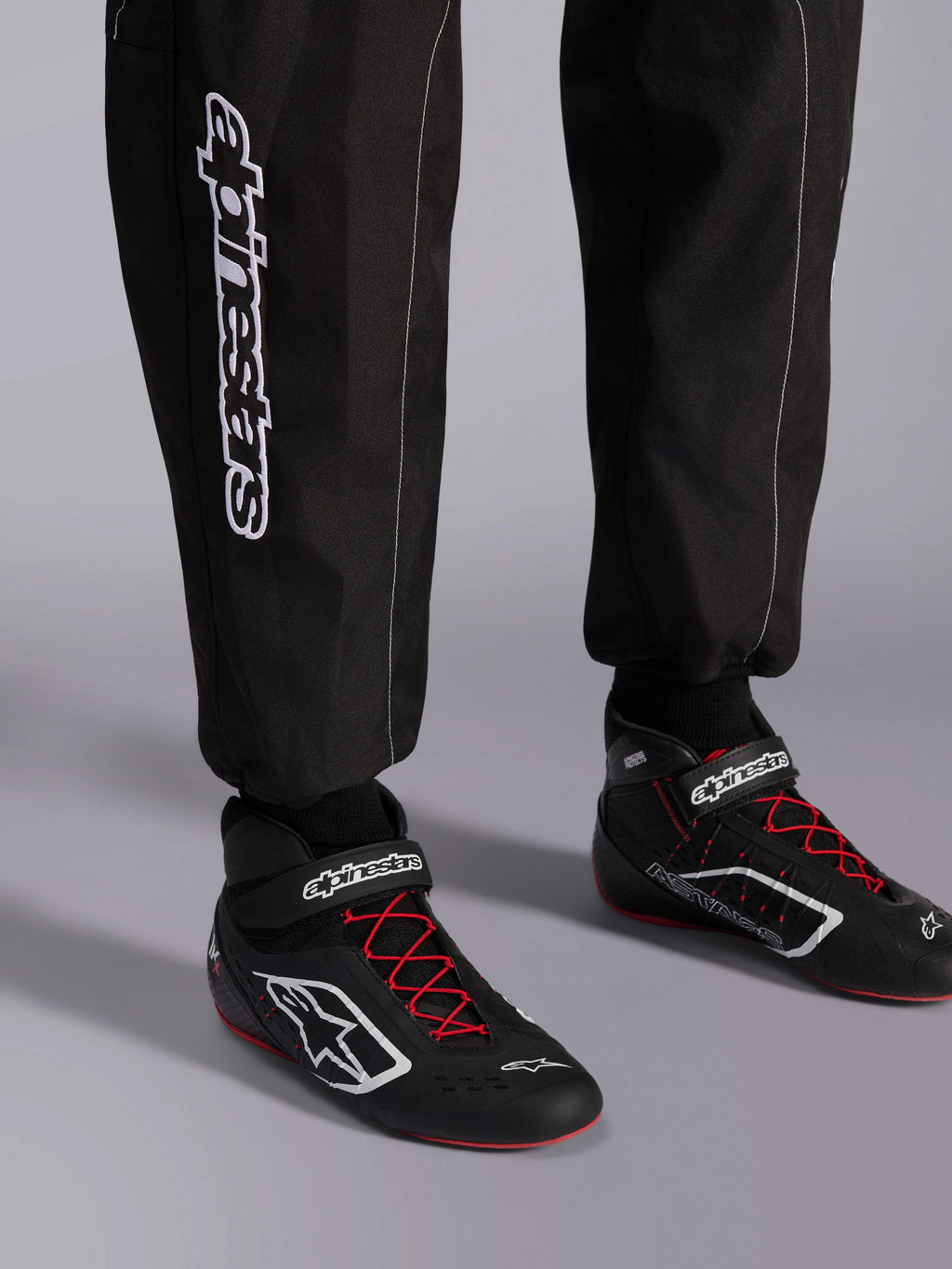 Alpinestars KMX-9 V3 Suit, mono de karting, negro, rojo y blanco, vista detallada de la parte inferior de las piernas y botas de karting, marca Alpinestars blanca en el lateral, calzado negro con cordones rojos y detalles blancos, equipamiento profesional de carreras