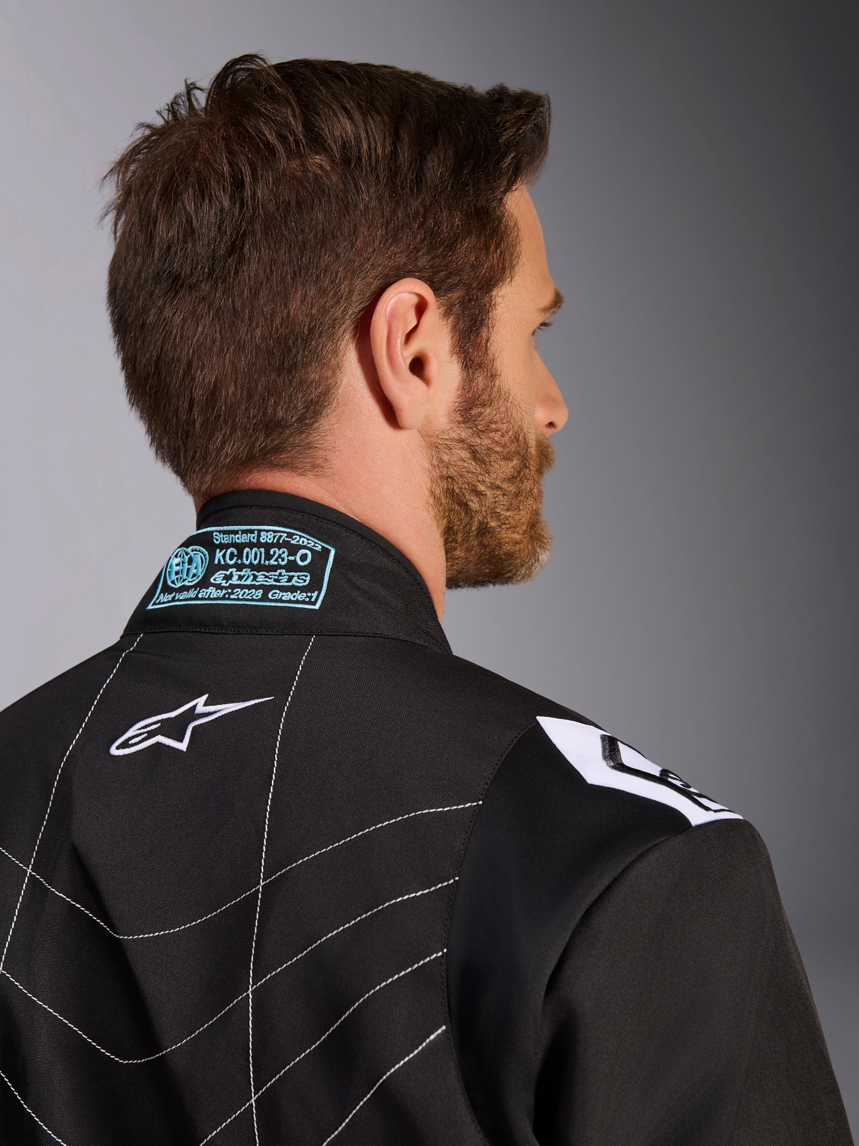Mono Alpinestars KMX-9 V3, negro y blanco, primer plano de la parte posterior del cuello y el hombro, con detalles de homologación FIA bordados en azul claro, logotipo de estrella blanca y costuras blancas en contraste.
