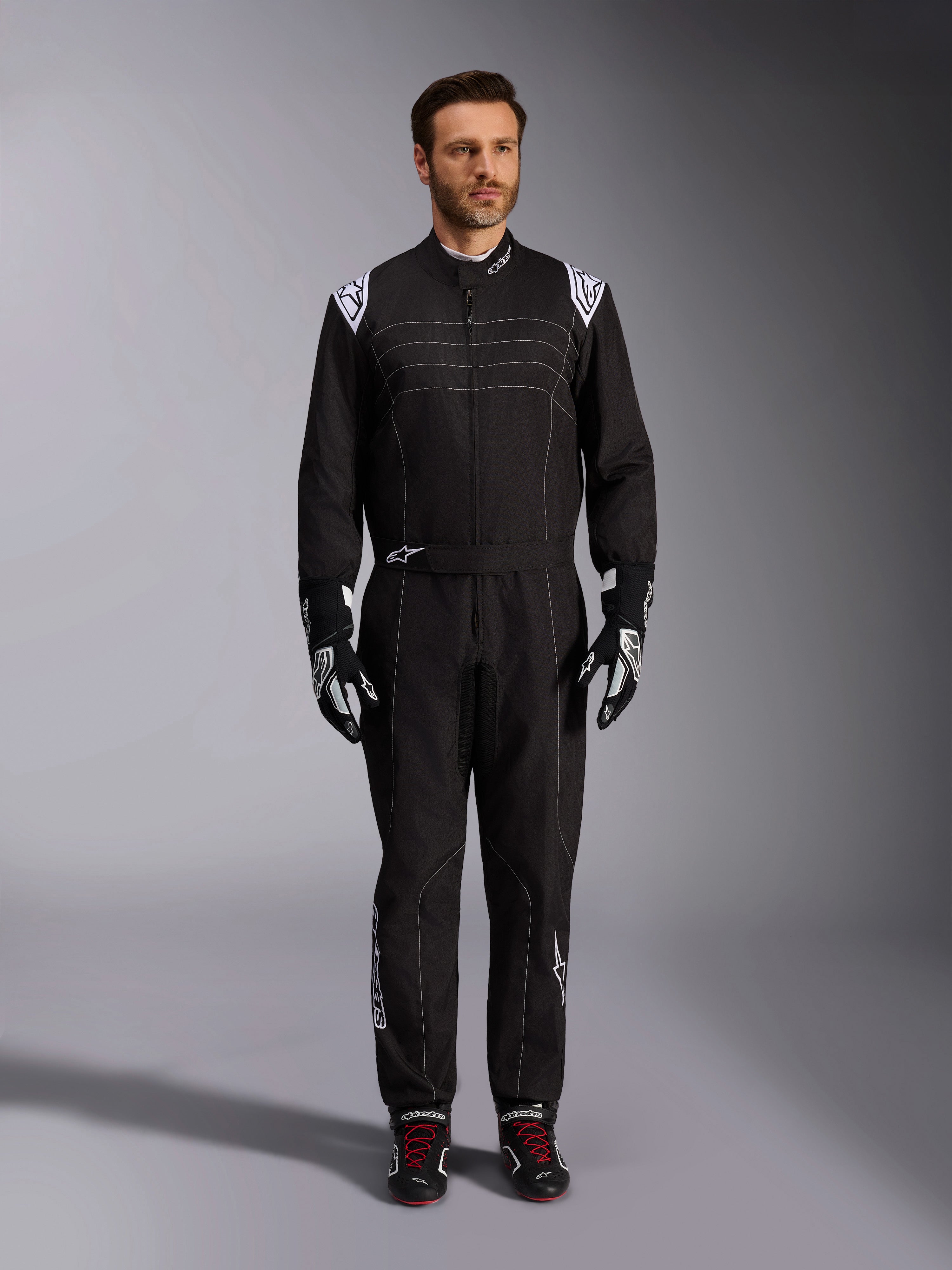 Mono Alpinestars KMX-9 V3, traje de karting negro y blanco, vista de cuerpo completo en un modelo, con logotipos y detalles blancos en los hombros, combinado con guantes de carreras en blanco y negro y calzado negro con cordones rojos, ajuste atlético-regular para equipamiento de karting profesional.